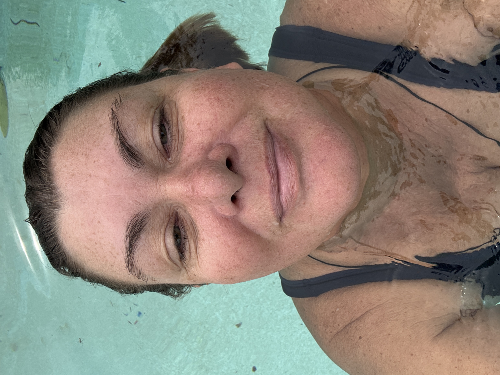 A close-up, overhead shot shows a woman submerged in clear, turquoise water, likely a swimming pool. Her face is the primary subject, with freckles visible across her nose and cheeks. Her eyes are partially closed, and she has a slight smile. She wears a dark-colored swimsuit. The water is calm, with some ripples and light reflections visible on the surface. Debris, such as small leaves or twigs, is scattered across the water in the background. The lighting suggests it is daytime. The location is identified as Baan Bang Rak, Thailand.