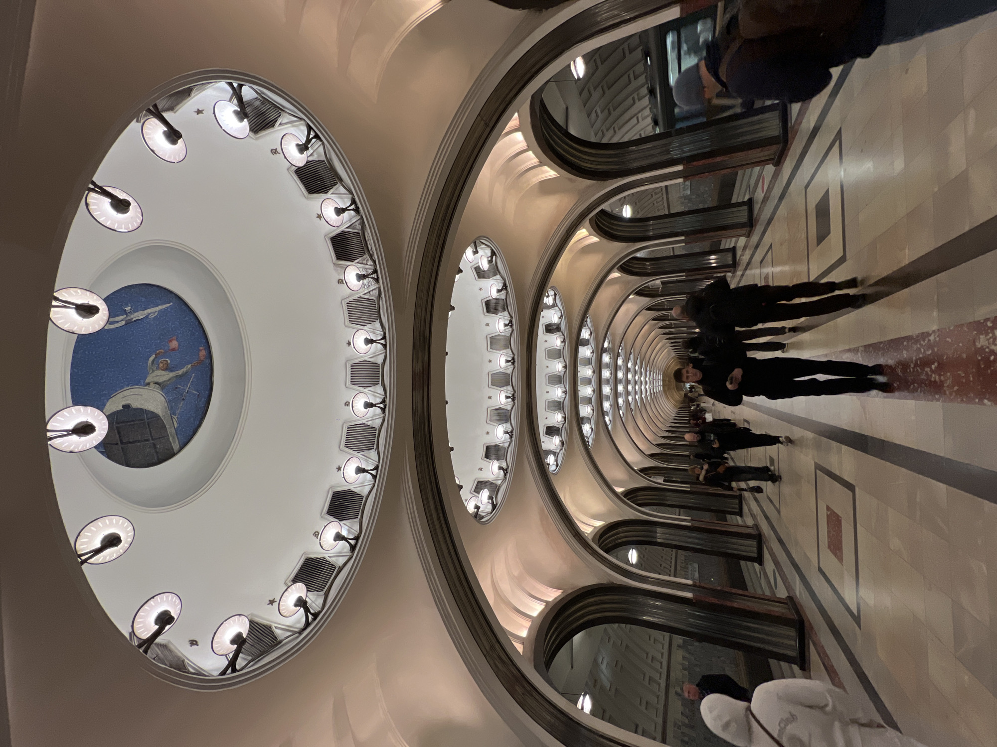The image depicts the interior of a Moscow Metro station, specifically the Komsomolskaya station, located on the Circle Line. The scene shows a long corridor with a series of arched openings on one side, leading to a parallel platform. The architecture is characterized by ornate, Stalinist-era designs, including rows of decorative arches, columns, and ceiling fixtures with lamps. The ceiling features intricate mosaic artworks and patterns. Several people are walking through the corridor, appearing as blurred figures due to movement. The floor is tiled with geometric patterns. The overall lighting is warm, creating a sense of depth and perspective.
