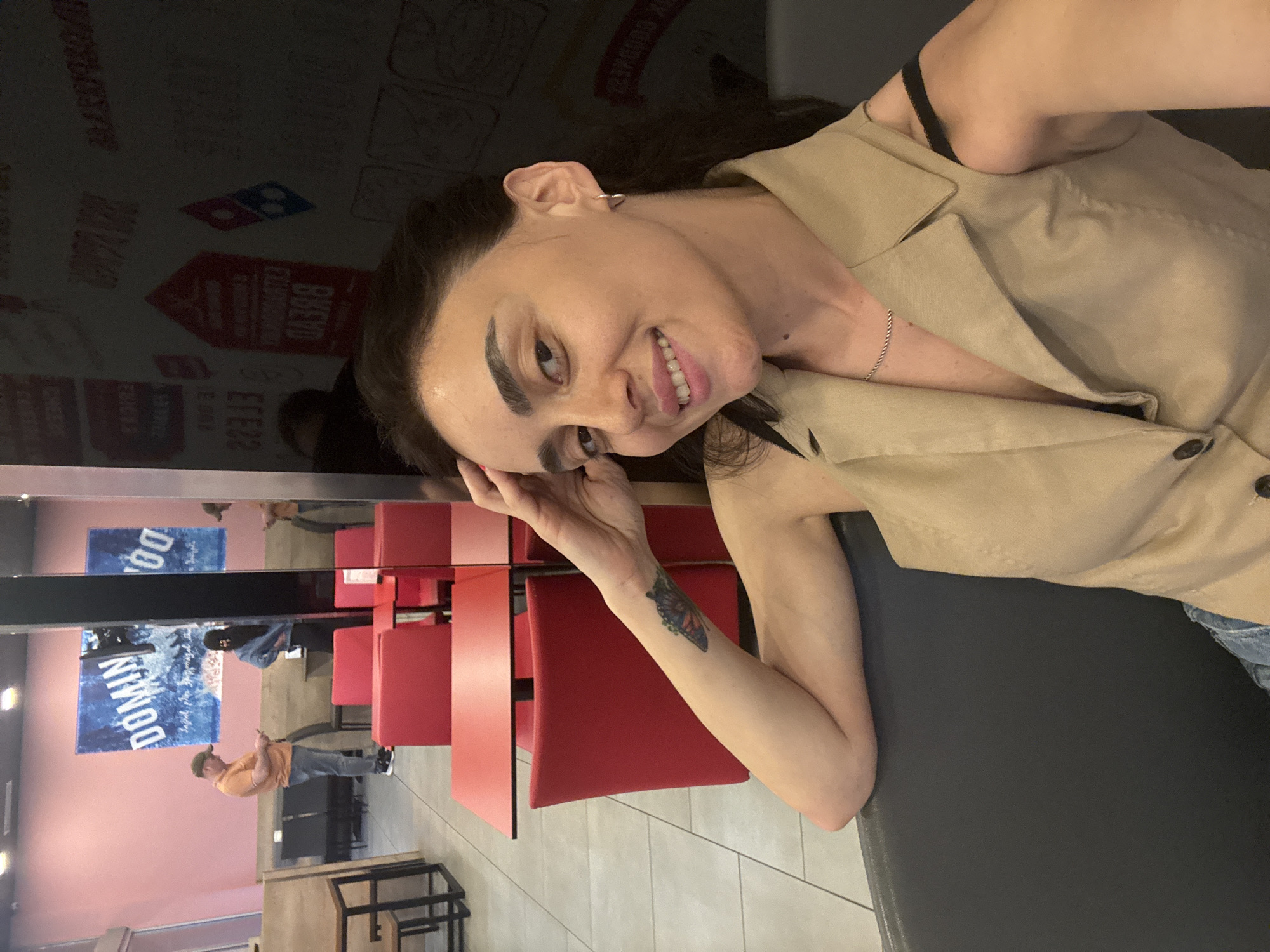 The image shows a young woman with dark hair and brown eyes, posing for a selfie. She is wearing a light-colored button-up shirt and denim jeans. A delicate silver chain necklace is visible around her neck. She has a small butterfly tattoo on her left forearm. Her facial expression is playful, with a slight smile and her head tilted to the side. The background is somewhat blurred but appears to be an indoor setting, possibly a restaurant, with red chairs and tables visible. In the reflection on the left, there are signs with the Domino's Pizza logo and text that appears to be in a foreign language, likely Georgian given the location context. The overall lighting suggests it might be daytime or well-lit indoor lighting.