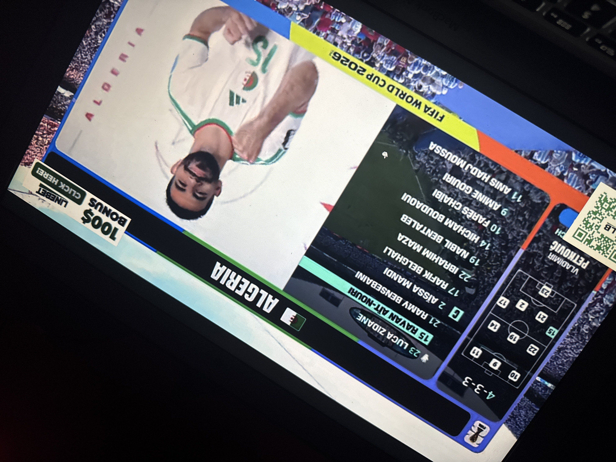 The image shows a laptop screen displaying a sports-related graphic. The graphic features a football formation diagram labeled "4-3-3" and a list of players with numbers, including "23 LUCA ZIDANE," "15 RAVAN AIT-NOURI," and "19 NABIL BENETALEB." The flag of Algeria is visible, along with the text "ALGERIA." A player in an Algerian jersey is prominently featured on the right side of the screen. There is also an advertisement banner at the top right corner that reads "100$ BONUS LINEBET CLICK HERE!". The bottom of the screen displays "FIFA WORLD CUP 2026". The scene is viewed from an angle, with the laptop on a dark surface, possibly a table, and a black keyboard is partially visible. The background of the graphic shows a crowd of spectators. There is no indication of the city of Kufena, Nigeria in the image.