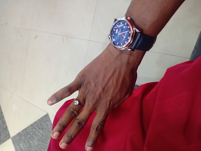 The image displays a close-up, high-angle view of a dark-skinned individual's left hand and forearm. A silver-colored ring with a prominent clear gemstone is worn on the fourth finger of the hand. On the left wrist, a watch with a rose gold-toned casing, a dark blue dial featuring multiple sub-dials and luminous markers, and a dark blue leather strap is visible. The individual wears a red garment, possibly a shirt or dress, exhibiting a subtle vertical striped texture. A sliver of a gray garment, likely trousers, is also discernible. The background consists of an indoor tiled floor, primarily light beige, with a section of dark gray and light beige checkerboard pattern in the upper left. The specific location within the city of Dorayi, Nigeria, cannot be determined from the visual information provided.