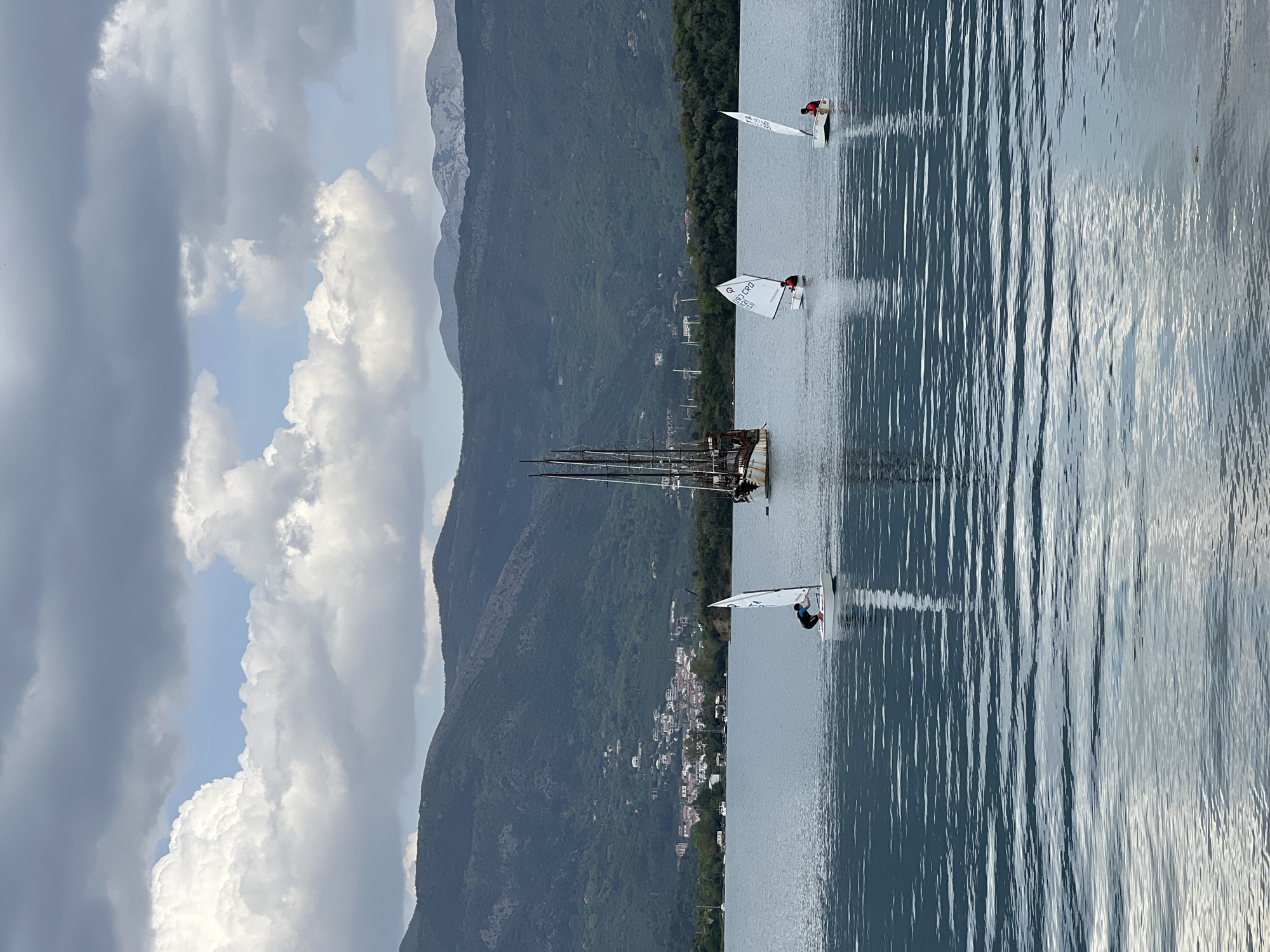 The image captures a picturesque coastal scene in Đuraševići, Montenegro, featuring sailing activities on calm waters with a mountainous backdrop.
In the foreground and midground, a tranquil body of water reflects the sky, with subtle ripples visible. Three small, white-sailed dinghies, likely Optimist class boats, are spread across the water. Each dinghy is occupied by a single person wearing a life vest, one red and one blue, actively engaged in sailing. Visible text on one sail reads "CRO 1603" and "P 135", while another clearly shows "CRO". Further left and closer to the shore, a larger, multi-masted wooden schooner or traditional sailing ship is anchored or moored, its intricate rigging a notable detail.
The background is dominated by steep, lush green, tree-covered mountains that rise dramatically. At the base of these mountains, along the shoreline, a scattering of white-roofed buildings indicates a small, charming coastal settlement or village.
The sky above features a mix of bright white cumulus clouds and darker grey clouds, interspersed with patches of clear blue sky, suggesting variable weather conditions, perhaps late morning or early afternoon. The overall atmosphere is one of peaceful recreational or training activity on the water.