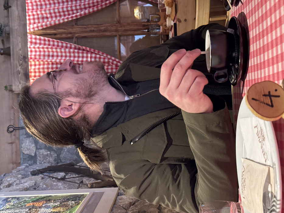 A male individual is seated at a table in what appears to be a rustic restaurant or cafe in Jabuka, Montenegro. He is positioned in profile, looking towards the right side of the frame with a smiling expression. The man has medium-length brown hair tied in a low ponytail and wears glasses with light frames. He has a light brown beard and mustache. His attire consists of an olive green jacket over a dark green zippered fleece. His right hand holds a white stirring stick in a dark brown/black coffee cup, which rests on a matching saucer on a red and white checkered tablecloth. A wooden table number "4." is visible on the table. To his left, a white plate holds a folded white paper napkin with "AMANET" branding and a clear drinking glass. The background features a stone wall on the left with a framed landscape artwork. On the right, a wooden-framed window is adorned with red and white checkered curtains, tied back. Above the window, a wooden beam holds small potted plants and a metal object. The overall environment suggests a warm, interior setting.