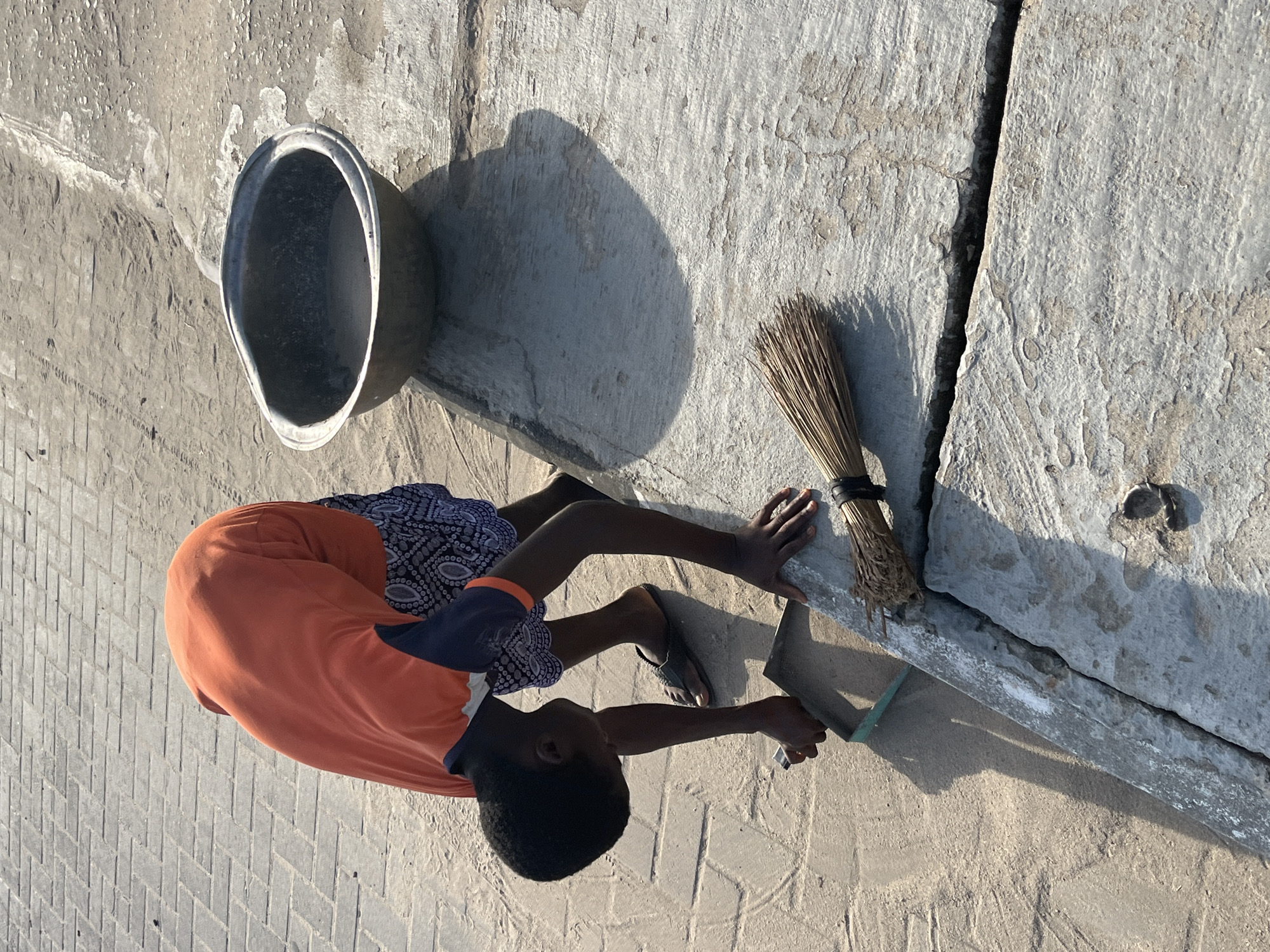 A young boy in an orange t-shirt is cleaning the edge of a concrete slab with a trowel in Badagry, Nigeria. Next to him is a bundle of sticks tied together, a metal bowl, and concrete slabs. The ground surface has a herringbone pattern. The image is taken from an overhead perspective.