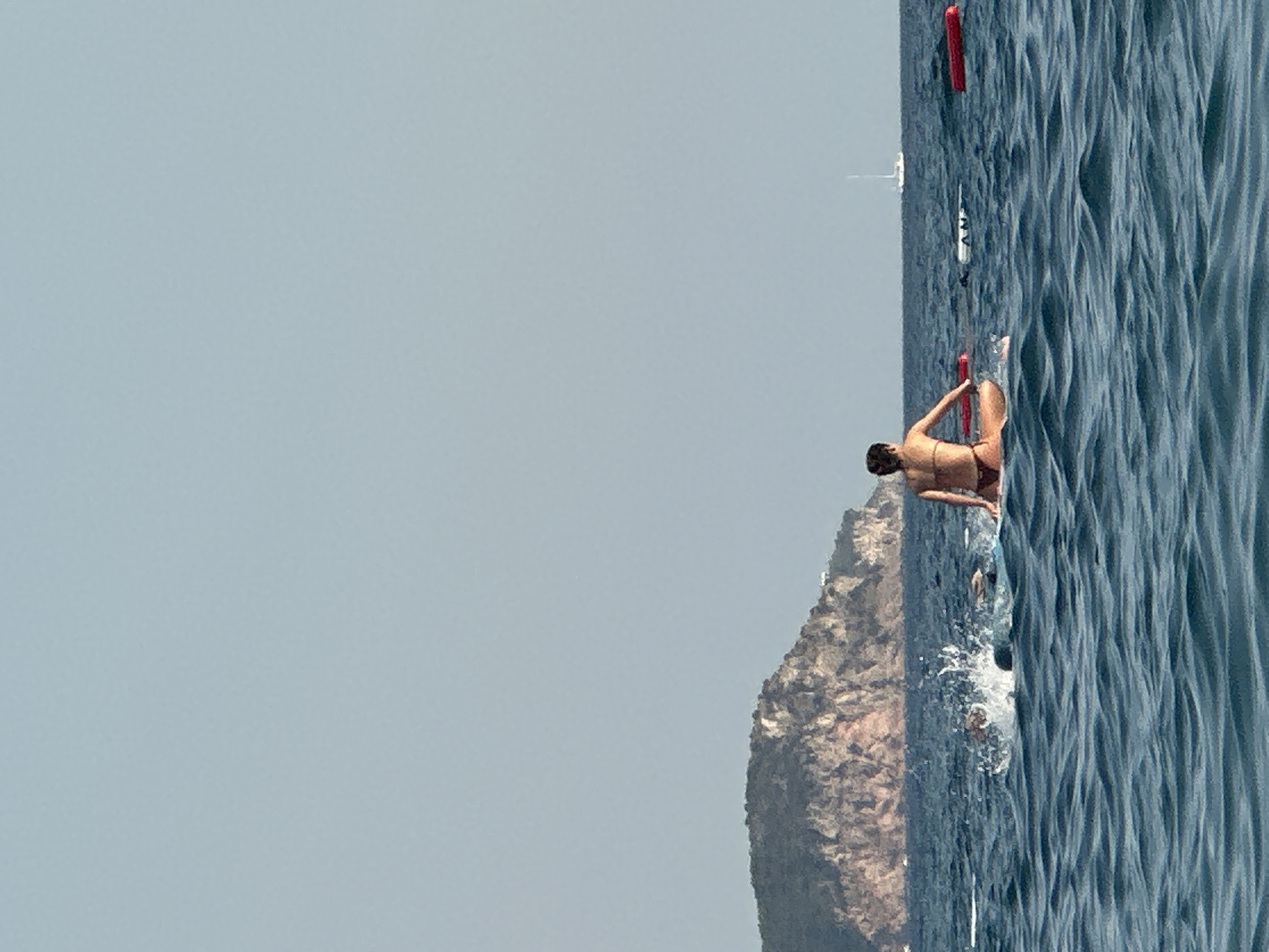 A woman in a bikini sits on a paddleboard in the Adriatic Sea, facing away from the viewer. She is in the foreground, with a mountainous island in the background. The sea is choppy with moderate waves. There are several red buoys floating in the water to the right of the woman. Further in the distance, a white sailboat is visible on the horizon. To the left of the woman, a person is swimming, creating a splash in the water. The sky is a clear, pale blue. The scene appears to be set in the coastal waters near Boreti, Montenegro.