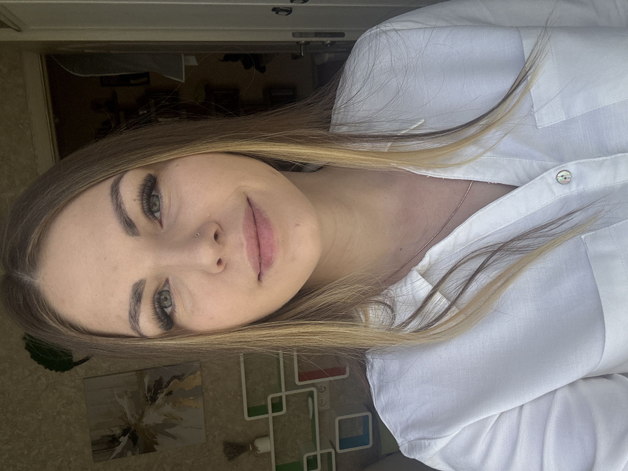 A woman is seen in a portrait oriented, close-up shot. She has fair skin, green eyes, and blonde hair with dark roots. She is wearing a white button-down shirt. A small nose ring is visible on her left nostril. The background includes a doorway, a painting on the wall, and a white shelving unit with colorful accents. The photo might have been taken in a room in Razdolie, Russia.