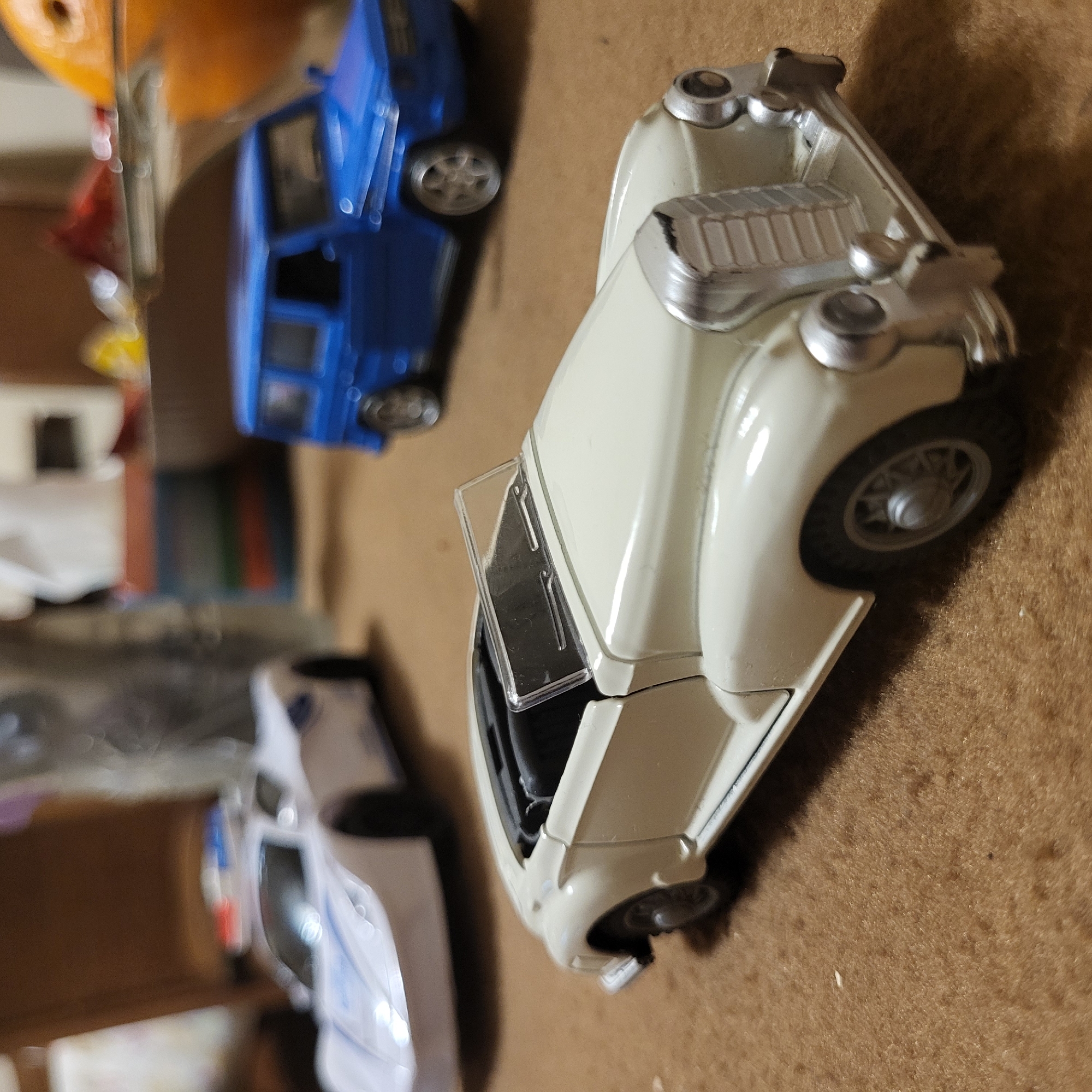 The image shows three toy cars on a brown surface, possibly a carpet or rug. The closest car is a cream-colored vintage model with an open driver's side door and visible engine detailing. Behind it is a blue toy car, followed by a blurry white car. The background is out of focus and shows a mix of colors and shapes. The location in Yerevan, Armenia, cannot be determined from the visual information provided.