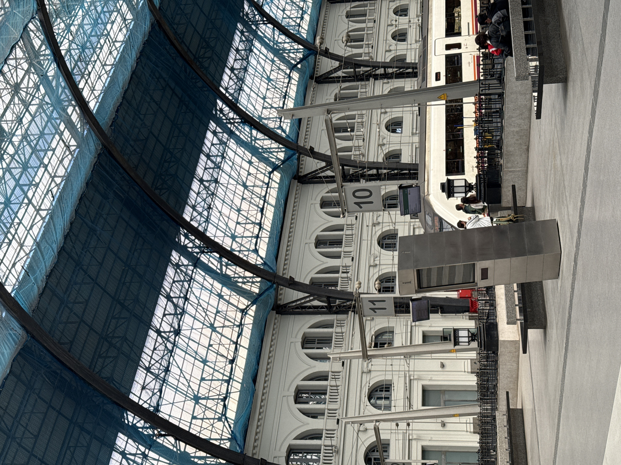 The image depicts the interior of a train station in Barcelona, Spain. The station features a high, arched glass and steel roof, with a blue safety net. The building's facade along the track is white and has rows of arched windows. There is a train parked at platform, and people are boarding and disembarking. Modern metal information displays and signage are visible. The ground is a gray concrete surface.