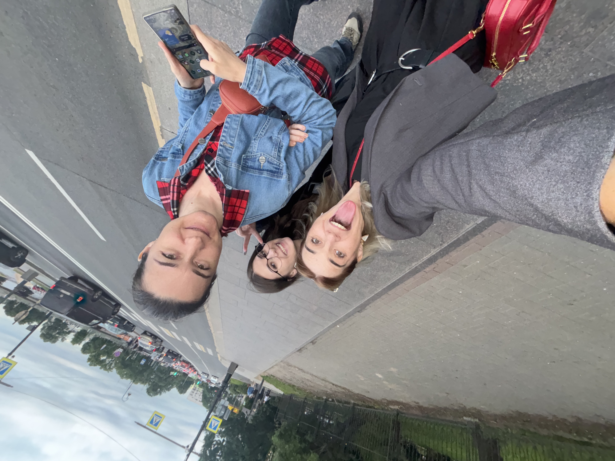 Three women are taking a selfie on a sidewalk in Saint Petersburg, Russia. The woman on the left is sticking her tongue out and wearing a grey blazer over a black top and pants with a red purse. The woman in the middle is smiling and wearing glasses. The woman on the right is holding a smartphone and wearing a denim jacket over a red plaid shirt. They are standing on a sidewalk next to a street with cars and pedestrian crossings. The sky is cloudy.