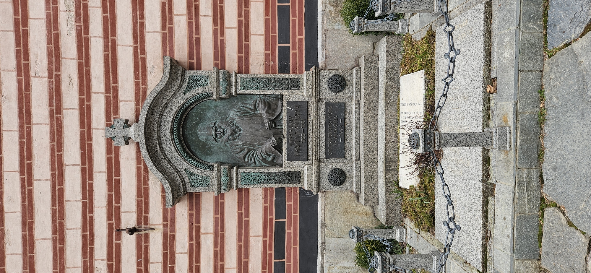 An outdoor commemorative monument for Hieromonk Neofit Rilski is depicted, situated against a multi-colored brick wall within the Rila Monastery complex in Rilski manastir, Bulgaria. The central feature is a verdigris-patinated bronze bas-relief of a bearded man in monastic robes and a skufia, holding a book, set within an arched, ornate granite frame topped with an Orthodox cross. Below the relief, a granite plaque is inscribed in Cyrillic, identifying the figure as "ЙЕРОМОНАХЪ НЕОФИТЪ РИЛСКІЙ" (Hieromonk Neofit Rilski) with the dates "1723–1881." A lower inscription reads "ОТЪ ПРИЗНАТЕЛНОТО УЧИТѢЛСТВО — 1908 —" (From the grateful teaching profession — 1908 —). The monument rests on a tiered stone base.
The background wall exhibits alternating horizontal courses of light-colored and red bricks, with a section of dark grey/black bricks near the monument's base. A single metal hook or bracket is visible on the left side of the wall. In the foreground, a low perimeter fence encloses the base of the monument, consisting of square-profiled granite posts with decorative caps, connected by dark metal chains. The ground within this enclosure features sparse moss and small, dry vegetation, along with a flat, light-colored stone slab. Beyond the fence, the ground is paved with irregular grey stone slabs. The scene is illuminated by ambient daylight, suggesting an overcast sky.