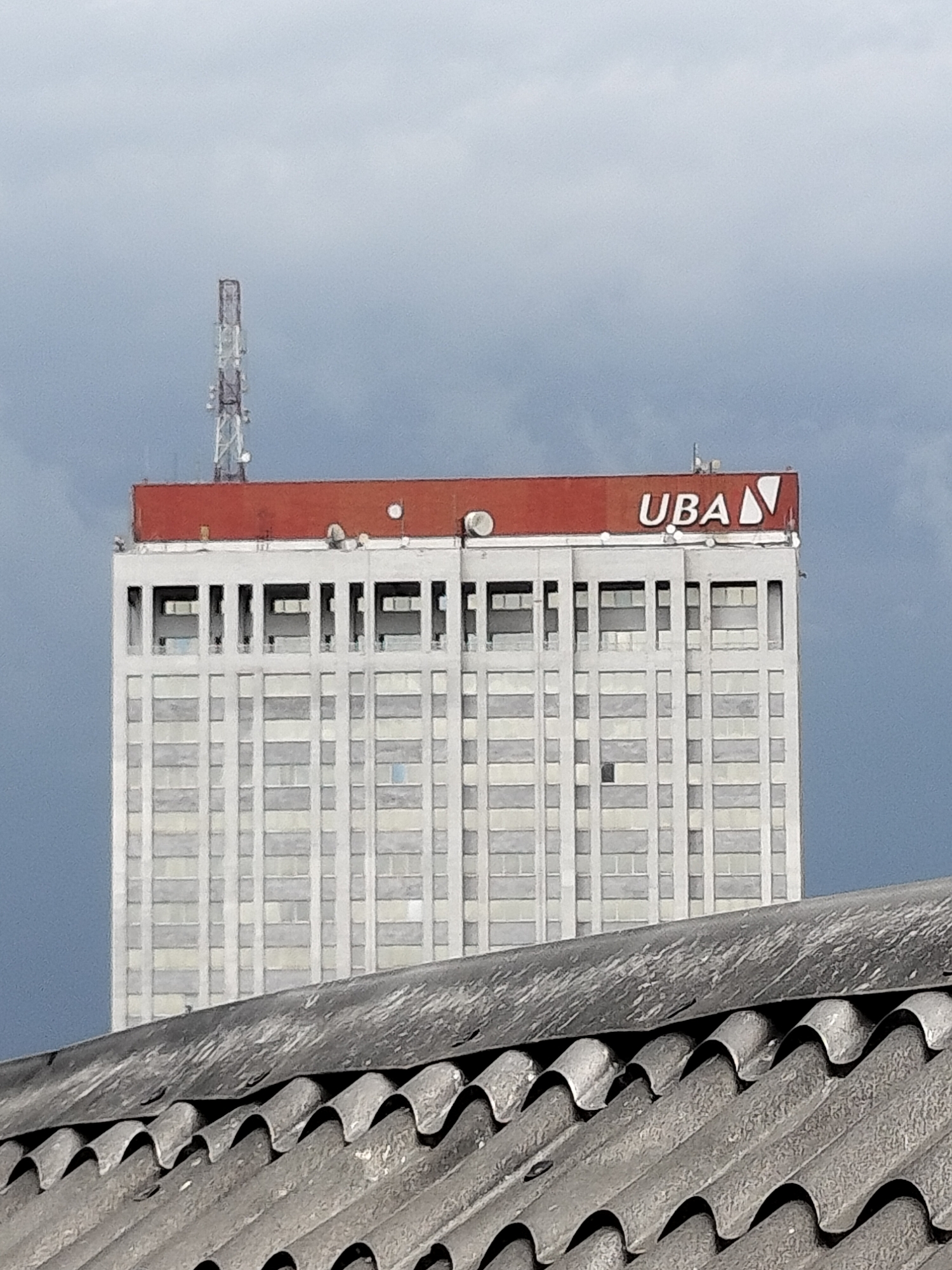The image shows the UBA House skyscraper in Lagos, Nigeria. The building is a tall, rectangular structure with a white facade and a red top. The UBA logo is prominently displayed on the top of the building. A communication tower is visible on the left side of the building's roof. The sky is overcast. In the foreground, there is a corrugated roof of another building.