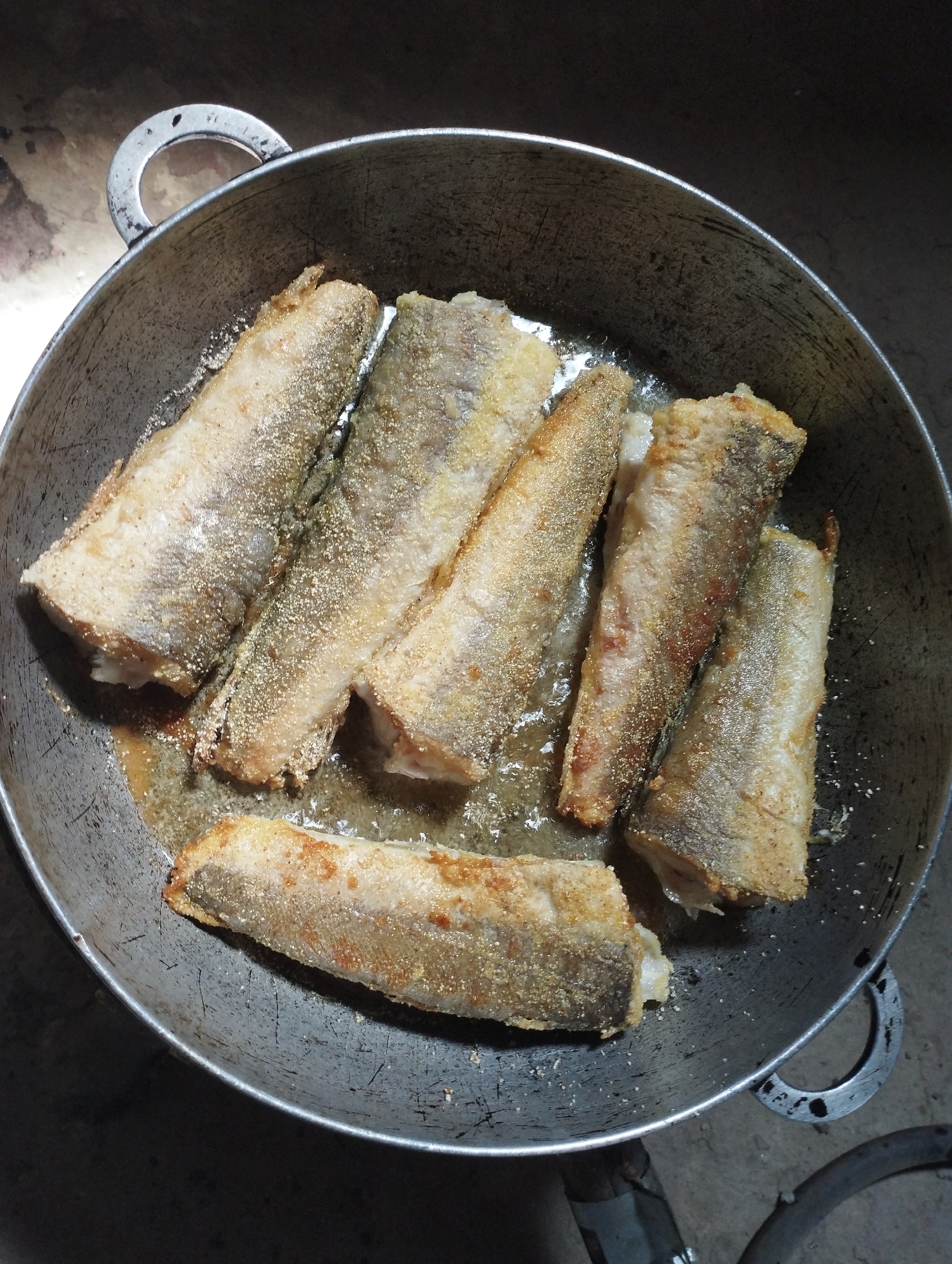 A close-up, overhead shot shows several pieces of breaded fish being fried in a dark, well-worn metal pot. The fish are golden brown and appear to be coated in a coarse meal or breadcrumbs. The pot is filled with oil, and some of the fish are partially submerged. The background is dark and indistinct, with a rough, textured surface visible at the top and bottom of the frame, suggesting an outdoor or rustic setting. The scene is likely in Fălești, Moldova, given the context provided, and depicts a simple, home-cooked meal in preparation.