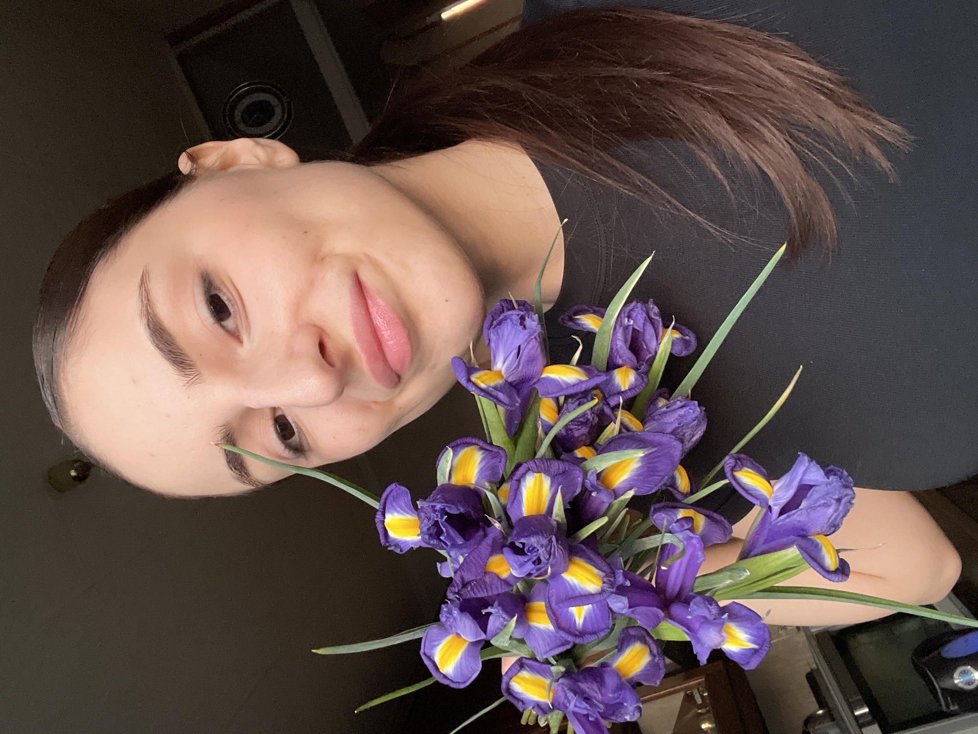 A young adult female is depicted in a self-portrait within an indoor residential setting in Moscow, Russia. She has dark brown hair styled in a ponytail, a fair complexion, and wears a black t-shirt. Her expression is a subtle smile, with direct eye contact towards the camera. She holds a compact bouquet of approximately 15 to 20 irises, characterized by deep purple petals with prominent yellow and white markings on the lower sepals, and visible green stems. The background is dimly lit and largely obscured, revealing a portion of what appears to be a kitchen area on the lower left, including a metallic object and a dark rectangular appliance. A circular, dark fixture is partially visible on the upper right against a dark ceiling.