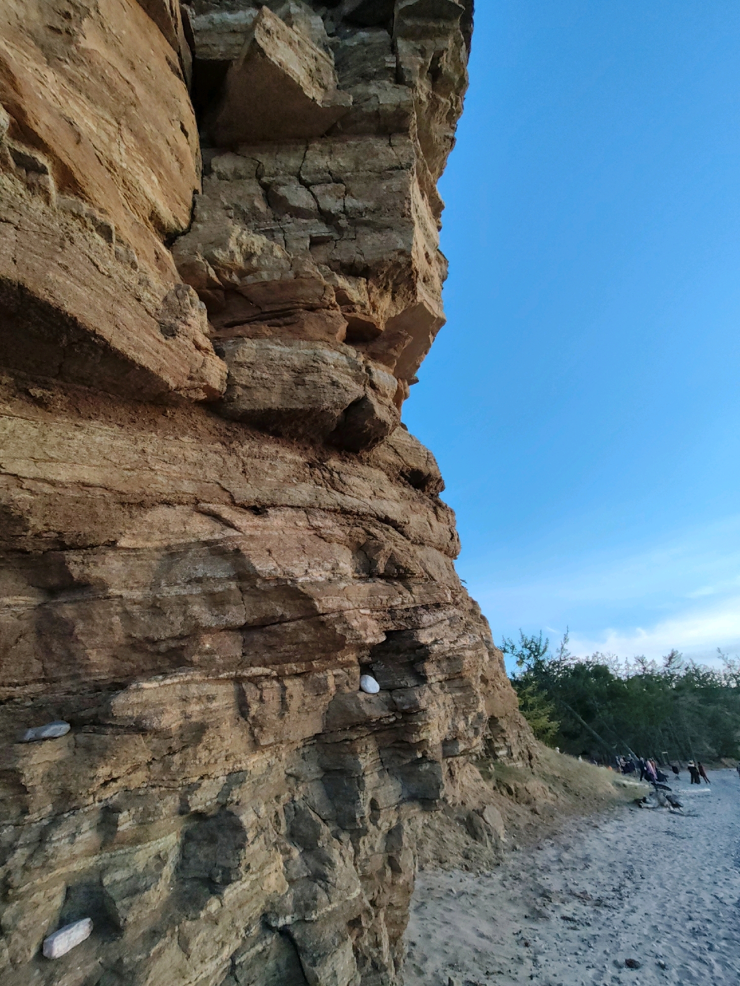 This is a low-angle, outdoor, full shot photograph taken at the Kharantsy, Russia location. The image features a large, layered, reddish-brown rock cliff face that dominates the left side of the frame. The rock shows visible strata and fissures, with a few small, white rocks embedded in its surface. To the right of the cliff, a clear, bright blue sky fills the upper portion of the image. Below the sky and extending to the right edge of the frame is a sandy beach with a few scattered pebbles. In the background, a line of dark green trees separates the beach from the sky. A small group of people, dressed in casual clothing, are visible on the beach in the mid-ground, appearing to be gathered or walking. The lighting suggests it is late afternoon or early evening, with the sun low in the sky.