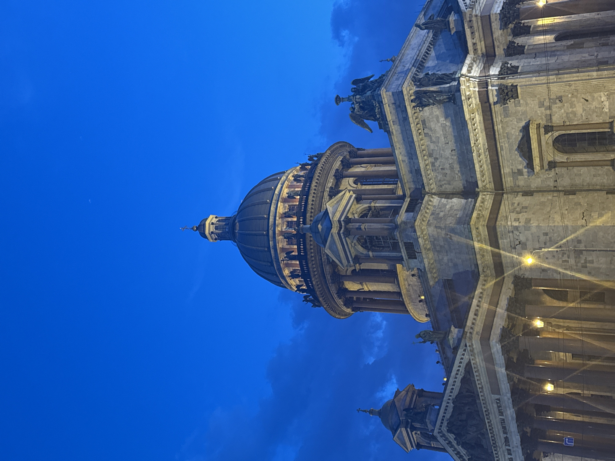 This media file captures a majestic architectural structure, likely a cathedral or grand public building, bathed in artificial light against a deep blue twilight or nighttime sky. The perspective is low, looking up towards the building's upper sections.
The central subject is a colossal building, constructed from light-colored stone or marble, exhibiting classical architectural elements. A prominent, dark-colored, ribbed dome, topped with a lantern and finial, dominates the upper left portion of the image. Surrounding the dome's base are numerous large columns, and more columns are visible along the building's main façade. The structure is richly adorned with intricate sculptures and statuary; several figures, including winged statues, are perched along cornices and rooflines. Parts of the detailed stone façade, featuring windows and decorative elements, are also visible. In the lower right, a section of another, smaller domed structure or a lower wing of the same building can be seen.
The activity depicted is static; it is an architectural photograph. The scene suggests evening or night, indicated by the deep indigo blue of the sky. Patches of darker, wispy clouds are scattered across the sky, particularly towards the right side of the frame. The building itself is illuminated by numerous hidden artificial lights, which cast warm, yellowish glows and create dramatic highlights and shadows on its textured stone surfaces. Some light sources appear as bright spots or starbursts on the lower parts of the building.
Visible notable details include Cyrillic text etched into a frieze along one of the building's cornices, though it's not fully legible from this perspective. The grand scale, classical design, and elaborate statuary are consistent with a significant landmark. The overall impression is one of grandeur and solemnity, captured under a dramatic night sky.