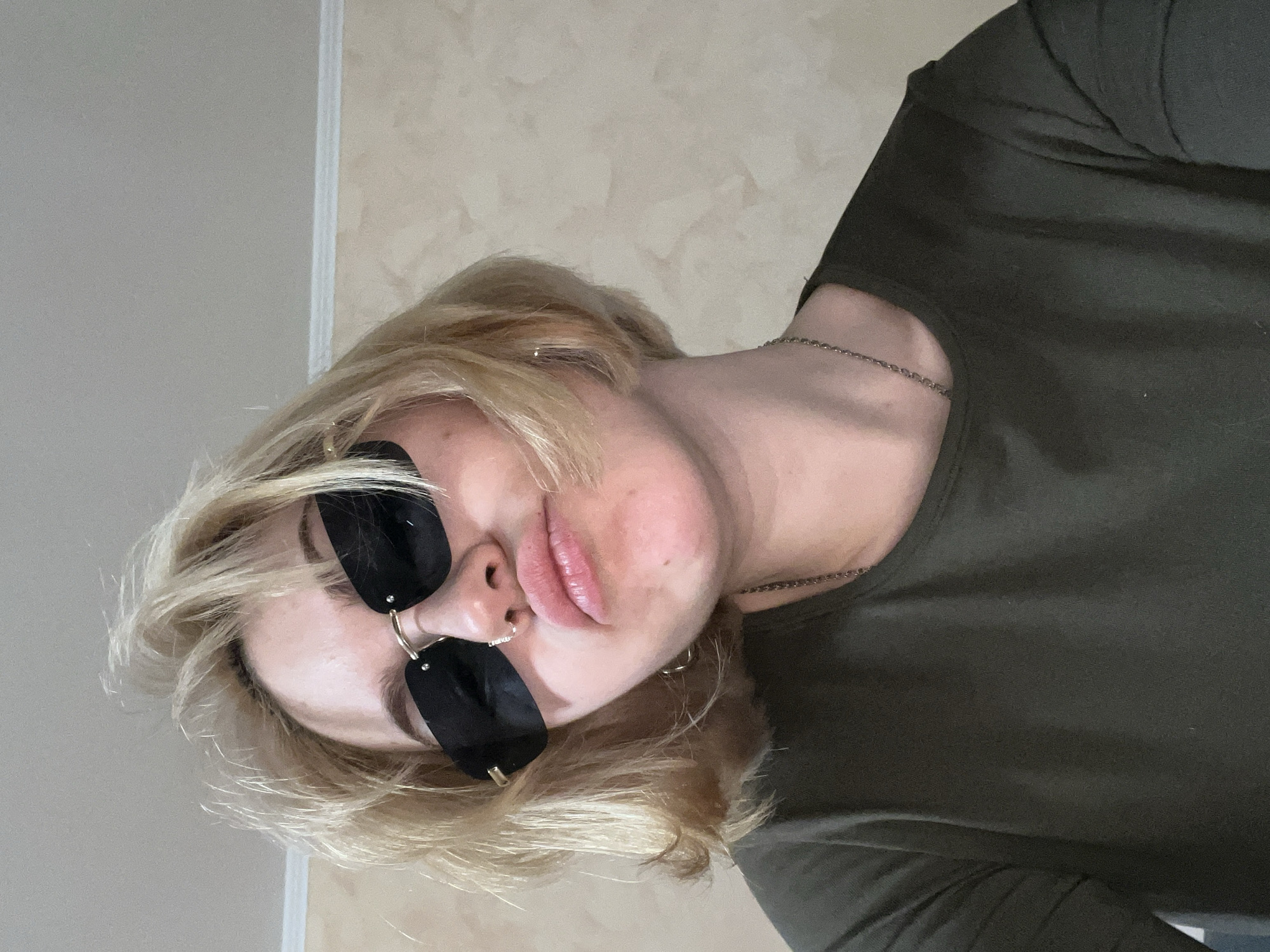 A medium close-up, slightly Dutch-angled shot features a person with short, wavy blonde hair. The individual is looking directly at the camera, wearing dark, rectangular sunglasses with gold-toned details near the nose bridge. A small, gold-toned hoop nose ring is visible on their right nostril. Their lips are full and slightly parted, revealing a natural pink hue.
The person is wearing a simple, dark olive green long-sleeved top, and a thin, silver-colored chain necklace is visible around their neck. The background consists of a plain, light-colored wall with a subtle textured pattern, suggesting an indoor setting. The lighting appears soft and even, possibly artificial. No text is visible in the image.