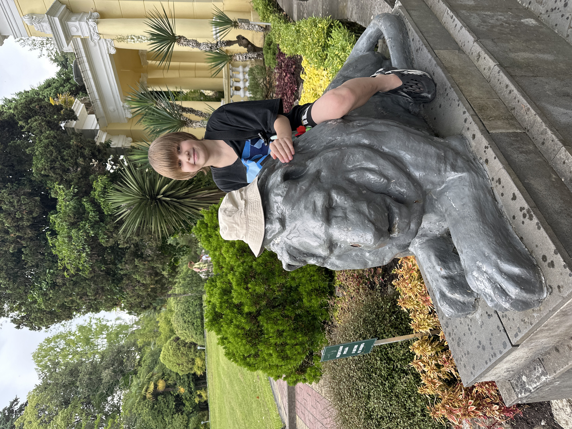 A young boy with blond hair and bangs, wearing a black t-shirt, black shorts with colorful accents, a light-colored bucket hat, and black shoes, sits atop a gray lion statue in a park. The boy is smiling at the camera. The lion statue is lying down on a stone pedestal. Behind the boy and the statue, there is lush green vegetation, including trees and bushes, and a yellow building with white columns. The scene appears to be outdoors in a park or garden. Based on the architectural style of the building and the surrounding vegetation, this location is likely the Dendrarium Botanical Garden in Sochi, Russia. A green sign with Russian text is visible to the left of the lion statue.
