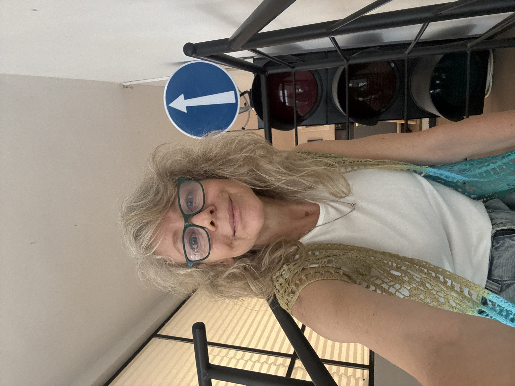 A woman wearing glasses, a white shirt, and a lace shawl appears to be indoors, possibly in Tel-Aviv, Israel, based on the presence of an arrow road sign with a single white arrow and a traffic light fixture with three circular lights. The background includes a metal structure, likely part of the indoor setting. The image suggests an artistic or quirky interior design element.