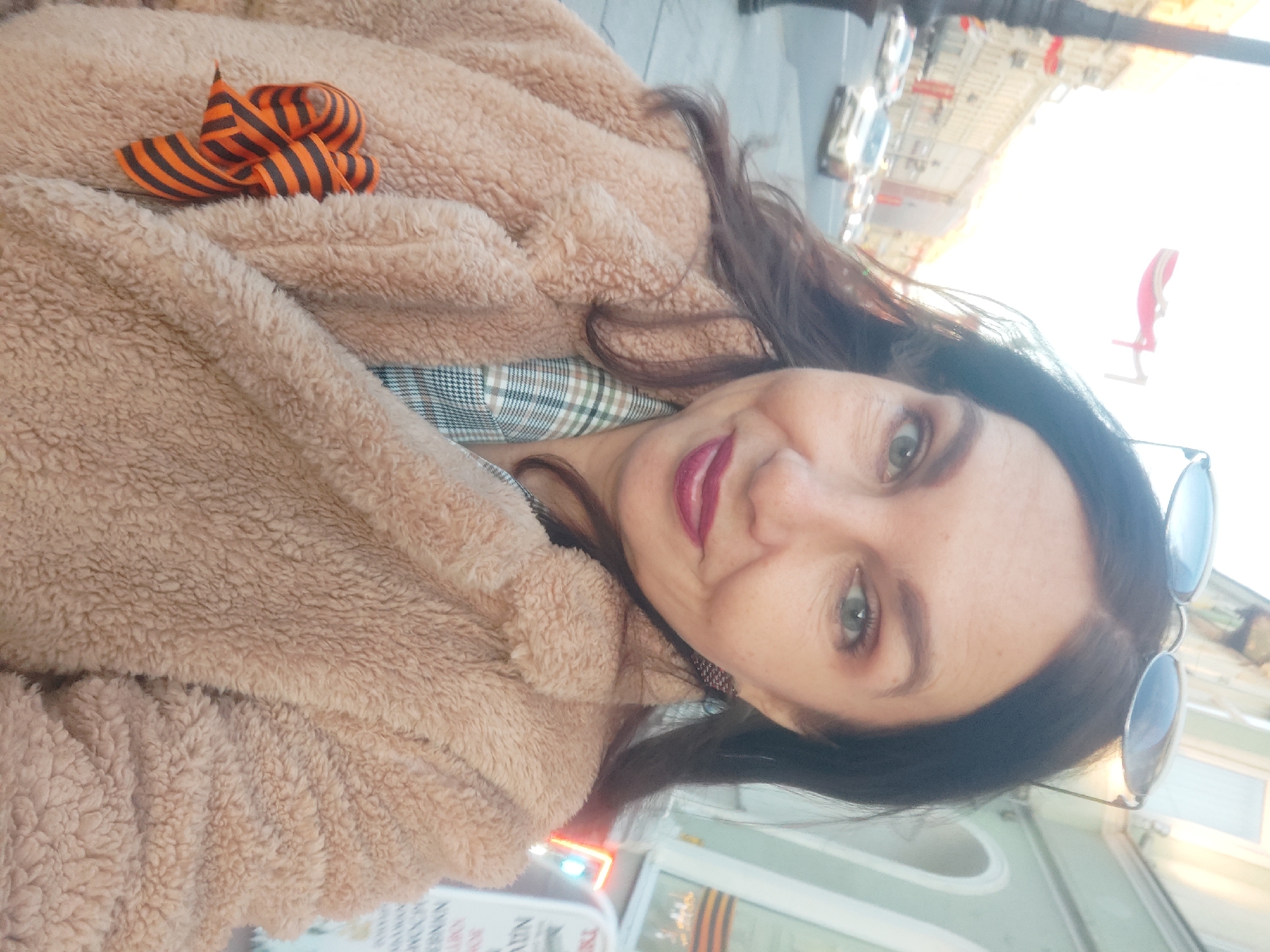 A middle-aged woman poses for a selfie in Saint Petersburg, Russia. She wears a light brown, fuzzy coat adorned with a St. George's ribbon. A plaid shirt is visible beneath the coat. She has dark hair, and sunglasses rest on her head. The background shows a city street with cars and a building archway.