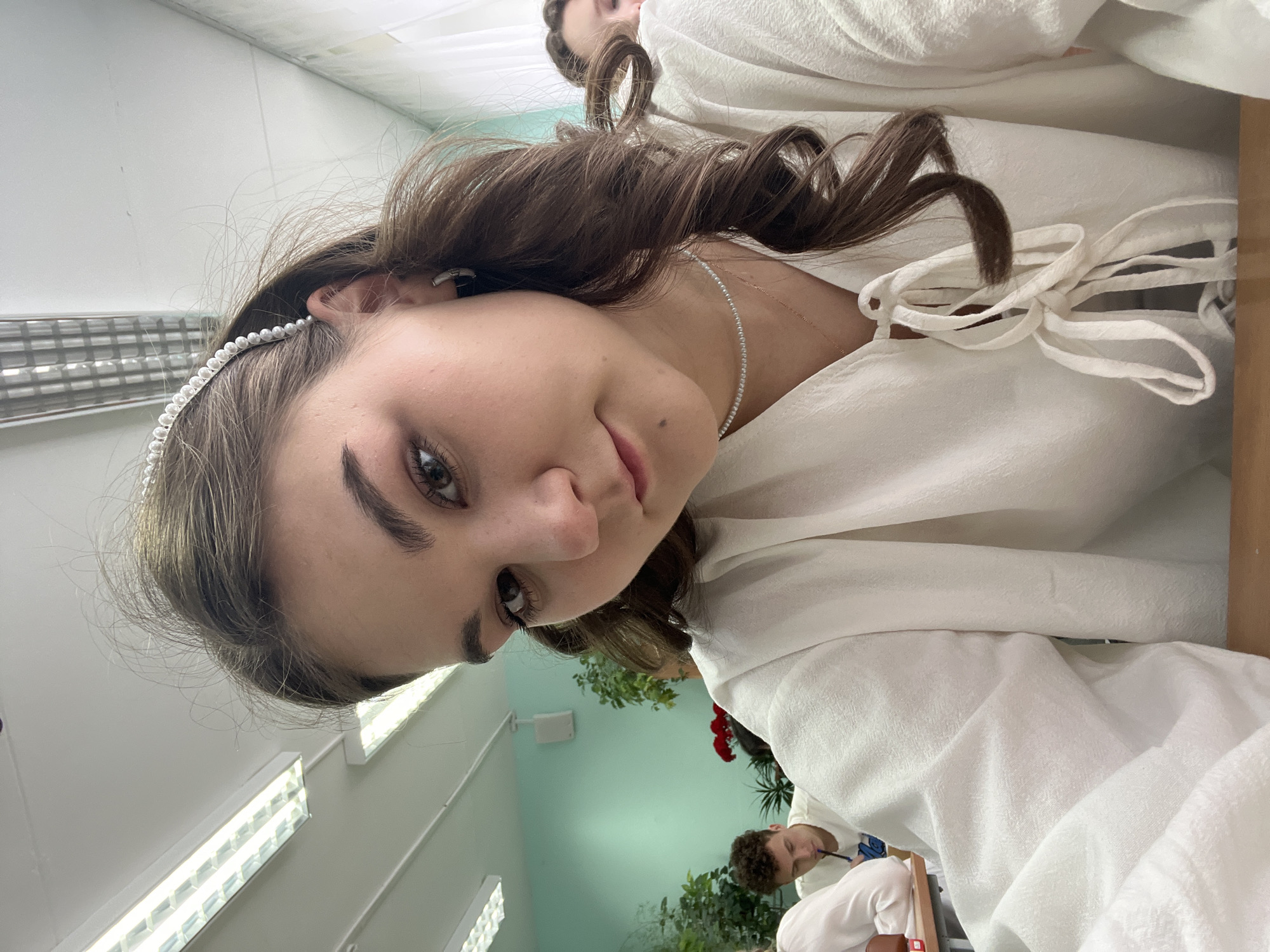 A low-angle close-up shows a young woman with brown hair styled in curls and wearing a pearl headband and a white linen-like blouse with tie details at the neckline. She is also wearing a delicate necklace. Her skin tone is fair, and her expression is relaxed. In the background, there are fluorescent lights on the ceiling, a light green wall, and some foliage, possibly potted plants. Other people are partially visible in the background, including a man wearing a white shirt. The setting appears to be indoors, possibly a classroom or office space. There is no clear evidence to confirm the location in Chaikovsky, Russia, but the style and context could be consistent with an educational or professional environment.