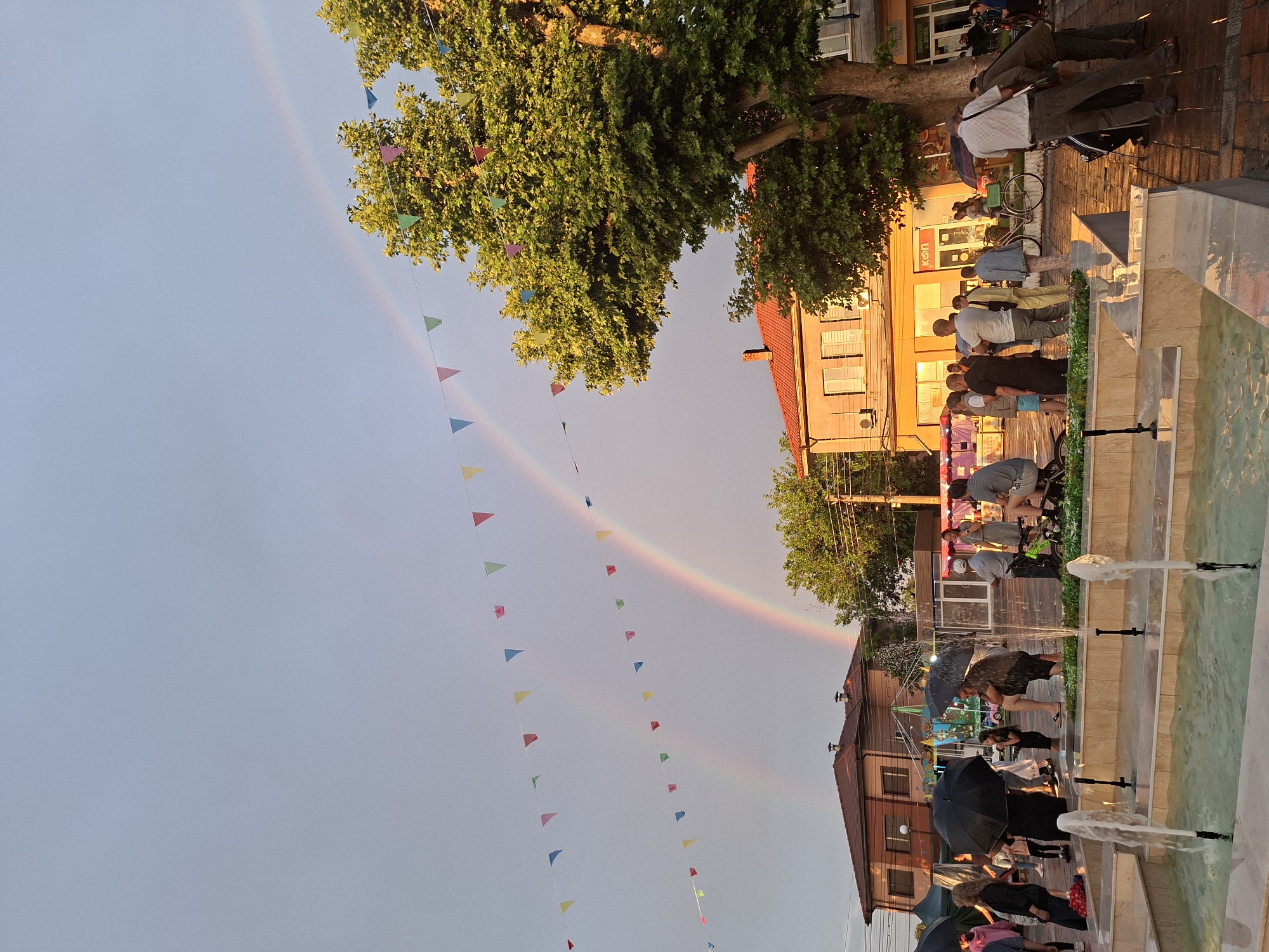 This image depicts a public square in Yasen, Bulgaria, during the late afternoon or early evening, shortly after a rain shower. A prominent double rainbow spans the sky, arching from the mid-left to the upper-right against a largely overcast grey-blue sky. Several strings of colorful triangular bunting flags, in various shades of red, yellow, blue, green, and pink, crisscross the upper half of the frame.
In the immediate foreground, a multi-tiered, light-colored stone fountain is actively spraying water from multiple jets. The ground surrounding the fountain and extending into the midground is paved, with visible wet patches indicating recent rain.
Approximately 20-30 individuals are dispersed throughout the midground. Many are holding open black umbrellas. Their attire is casual, consisting of t-shirts, shorts, and light pants. People are standing, conversing, and moving through the square. Notable individuals include a man in dark clothing squatting or kneeling near a market stall, a child nearby, and a man in a striped shirt leaning against a dark green bicycle on the far right.
To the left and center-right of the midground, various market stalls or tents are visible, one with a green covering and others with red and white awnings. A large, mature tree with dense green foliage occupies the right side of the frame, its leaves brightly illuminated by the low-angle sun.
In the background, two-story buildings are present. On the left, a building with a reddish-brown tiled roof and light-colored walls is partially visible. In the center-right, a building with a red-tiled roof and sun-drenched, light-colored walls features multiple windows and a commercial ground floor with a partially visible "K_N" sign. Utility poles and power lines are visible above the buildings and tree. The overall scene suggests a gathering or market event in a post-rain, festive atmosphere.