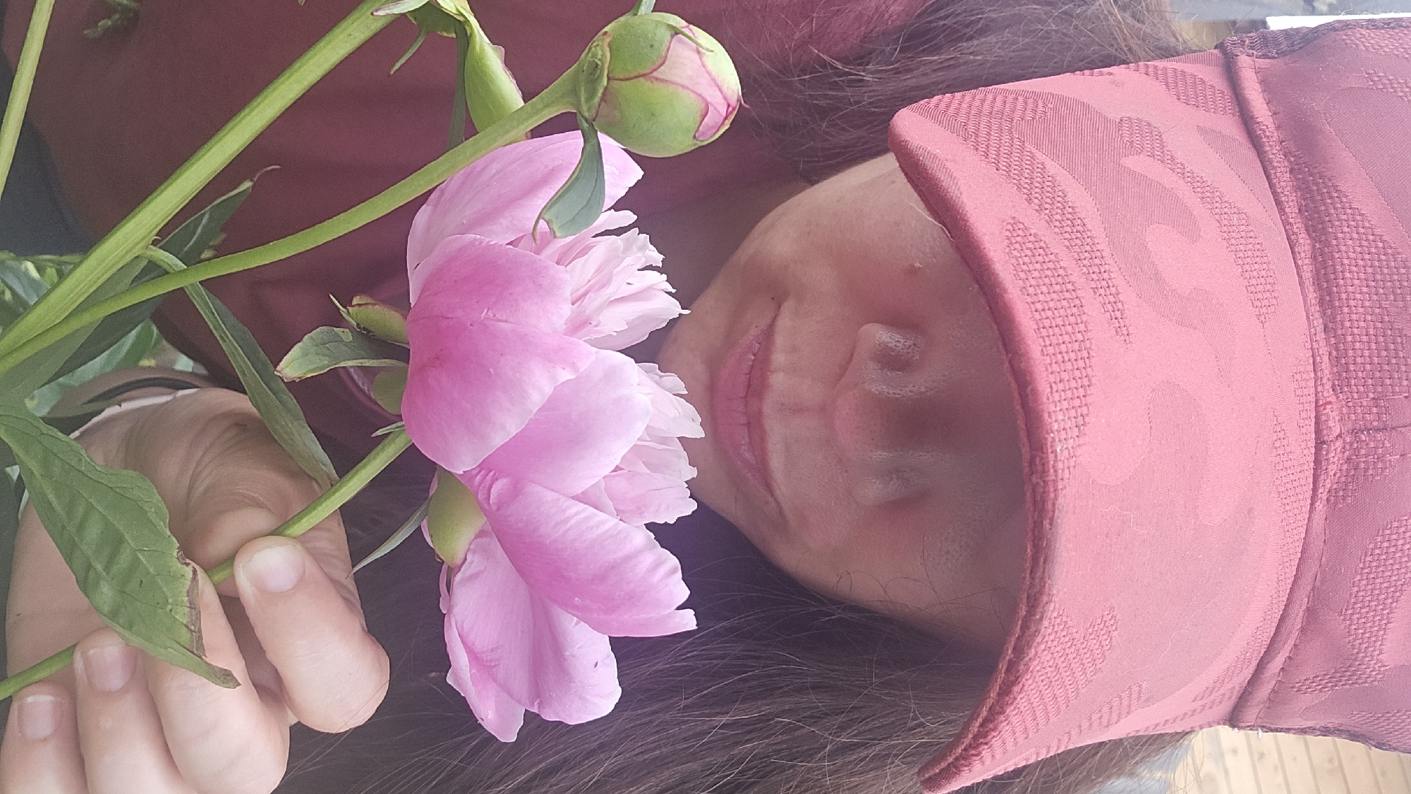 A close-up, low-angle shot shows a person wearing a red baseball cap and a maroon shirt, holding a pink peony flower and a bud. The person's face is partially obscured by the cap and the flower, with only their nose and lips visible. The background consists of a wooden wall, suggesting an outdoor setting, possibly in the city of Ermolaevo, Russia, given the provided context. The focus is on the flower and the person's hand, which is gently holding the stem of the peony. The lighting is soft, with diffused daylight illuminating the scene.