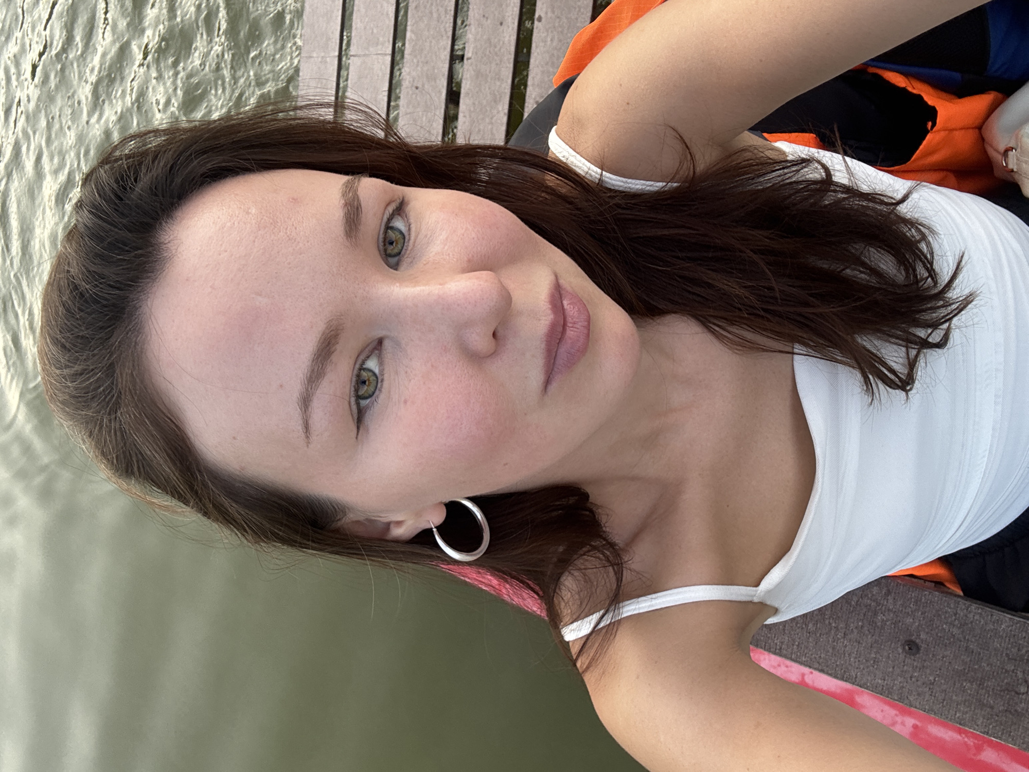 A woman with long, dark brown hair and green eyes is taking a selfie outdoors. She is wearing a white tank top and large silver hoop earrings. Her skin is fair, and she appears to be wearing light makeup. Behind her, there is calm water, and to her right, a wooden deck or pier. The woman is seated, with her arm visible in the foreground, and part of what appears to be an orange life vest is also visible. The image is taken from a slightly high angle. The setting suggests a waterside location, potentially a park or recreational area within the city of Ivanovo, Russia, given the context.