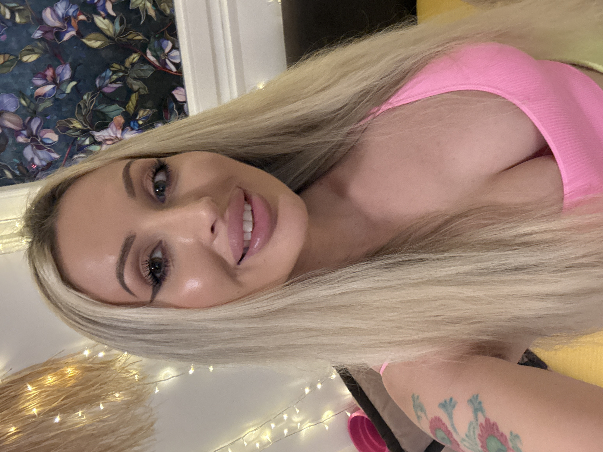 A medium shot photo shows a woman with long blonde hair and fair skin wearing a pink top. She is indoors, with decorative lights strung up behind her to the left, and a floral tapestry on the wall to the right. The woman has on makeup, including winged eyeliner and lip gloss. She is smiling, with her teeth visible. A tattoo is visible on her left arm. The background also shows some furniture and a hint of a yellow surface.
