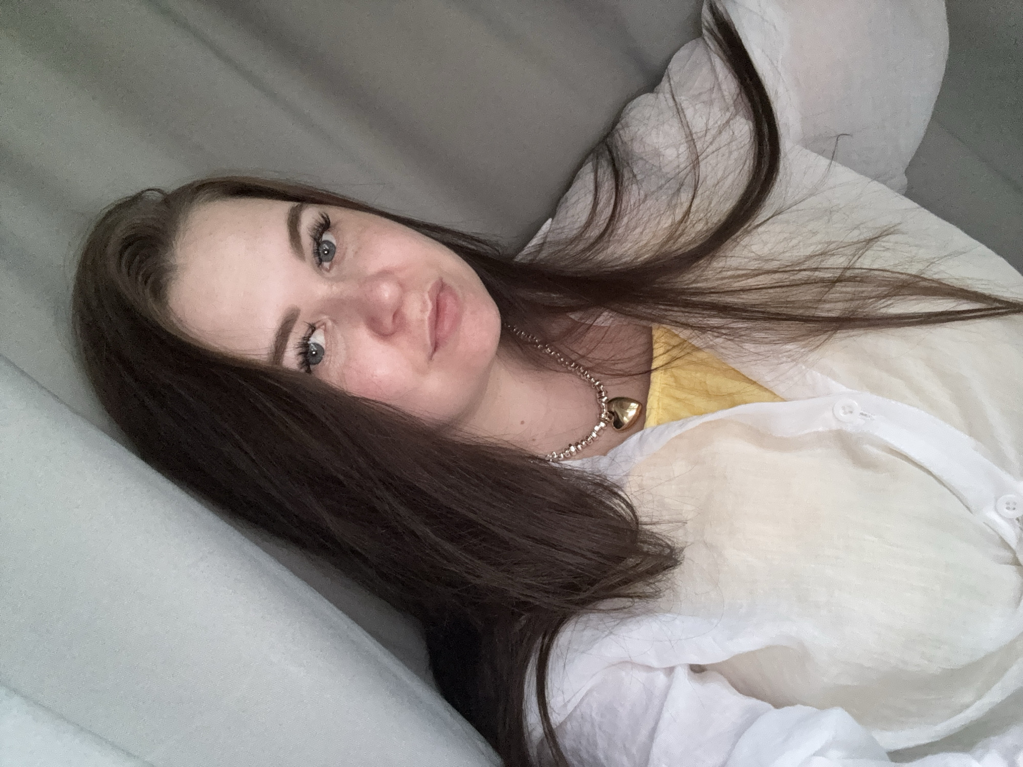 A close-up, head-and-shoulders selfie of a young Caucasian woman with long, dark brown hair and blue eyes is shown. She is wearing a yellow top underneath a sheer white open-collared shirt. A gold heart-shaped locket on a chain necklace is visible around her neck. The background appears to be a draped fabric, possibly curtains, in muted gray and light blue tones. The lighting suggests an indoor setting, likely a home, with no discernible elements placing the scene specifically in Chaikovsky, Russia. The woman is looking directly at the camera with a neutral expression.