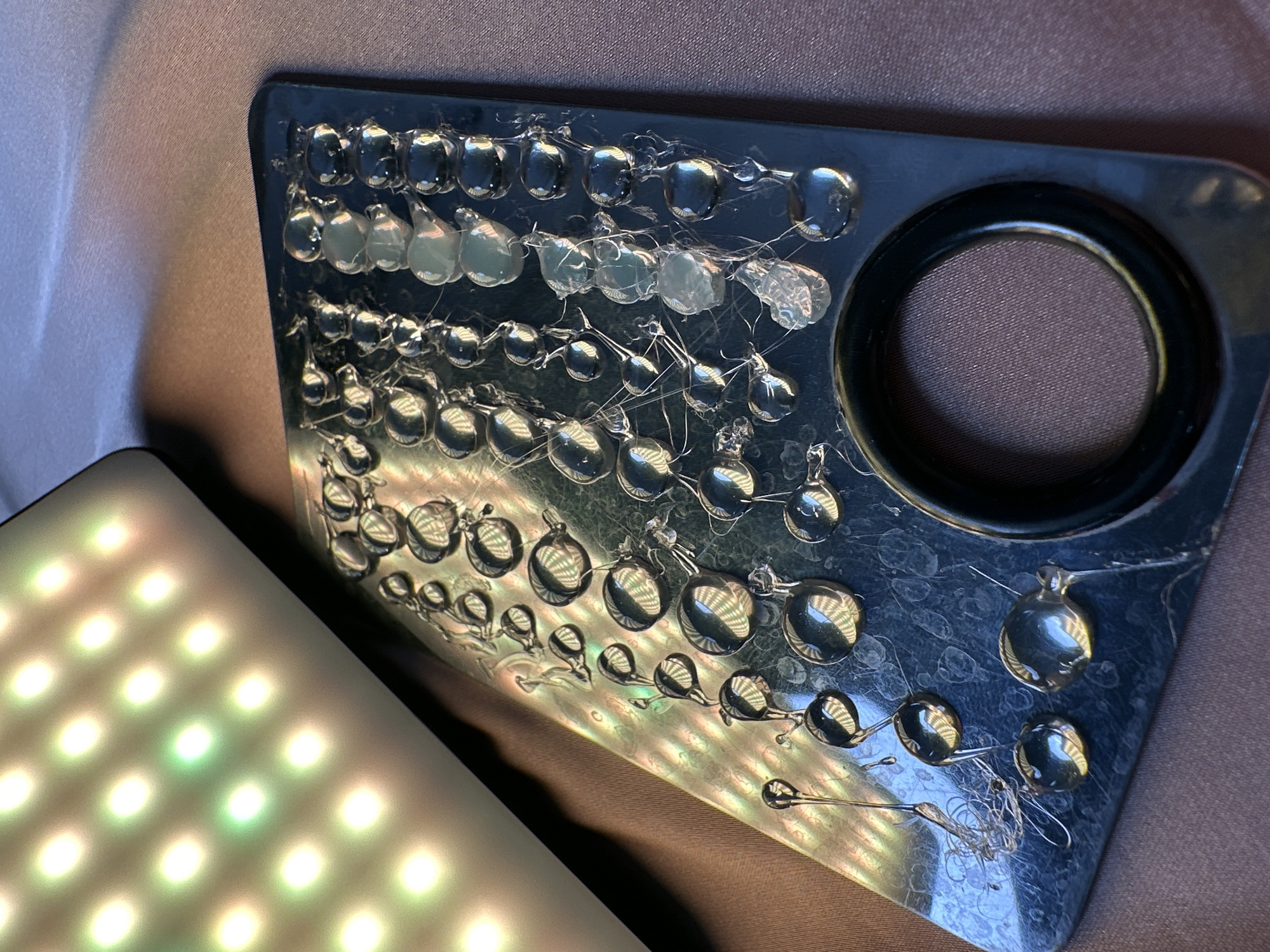 The image presents a close-up view of a dark, rectangular panel, possibly a rigid board, covered with numerous transparent, droplet-shaped formations. These forms, resembling dried hot glue, are varied in size and are often interconnected by thin, string-like filaments. Some droplets are clear and reflective, showcasing distorted grid patterns from an external light source, while others appear more opaque or cloudy. The surface of the dark panel itself has a slight sheen, exhibiting faint iridescent reflections in certain areas.
In the upper right corner of the panel, there is a large, circular opening that reveals a dark brown, textured fabric underneath. To the bottom left of the frame, a portion of a bright, illuminated light panel is visible. This panel displays a grid of square lights, predominantly emitting white and green hues, which appear diffused. The light emitted from this panel is visibly reflected in many of the transparent droplets on the dark panel. The background consists of a soft, brownish-purple fabric or surface. The scene suggests a crafting project, an experiment involving adhesives, or a study of light interaction with textured surfaces. There is no visible text. The location context provided is North Kuta, Indonesia.