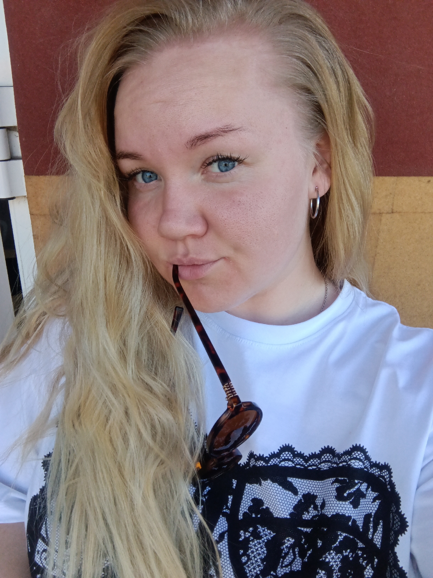 A blonde-haired woman with blue eyes is posing for a selfie. She is wearing a white t-shirt with a black lace design. She has sunglasses in her mouth and a silver hoop earring. The background is a combination of red, brown, and white. The city of Фоки, Russia, is not discernible from the image.