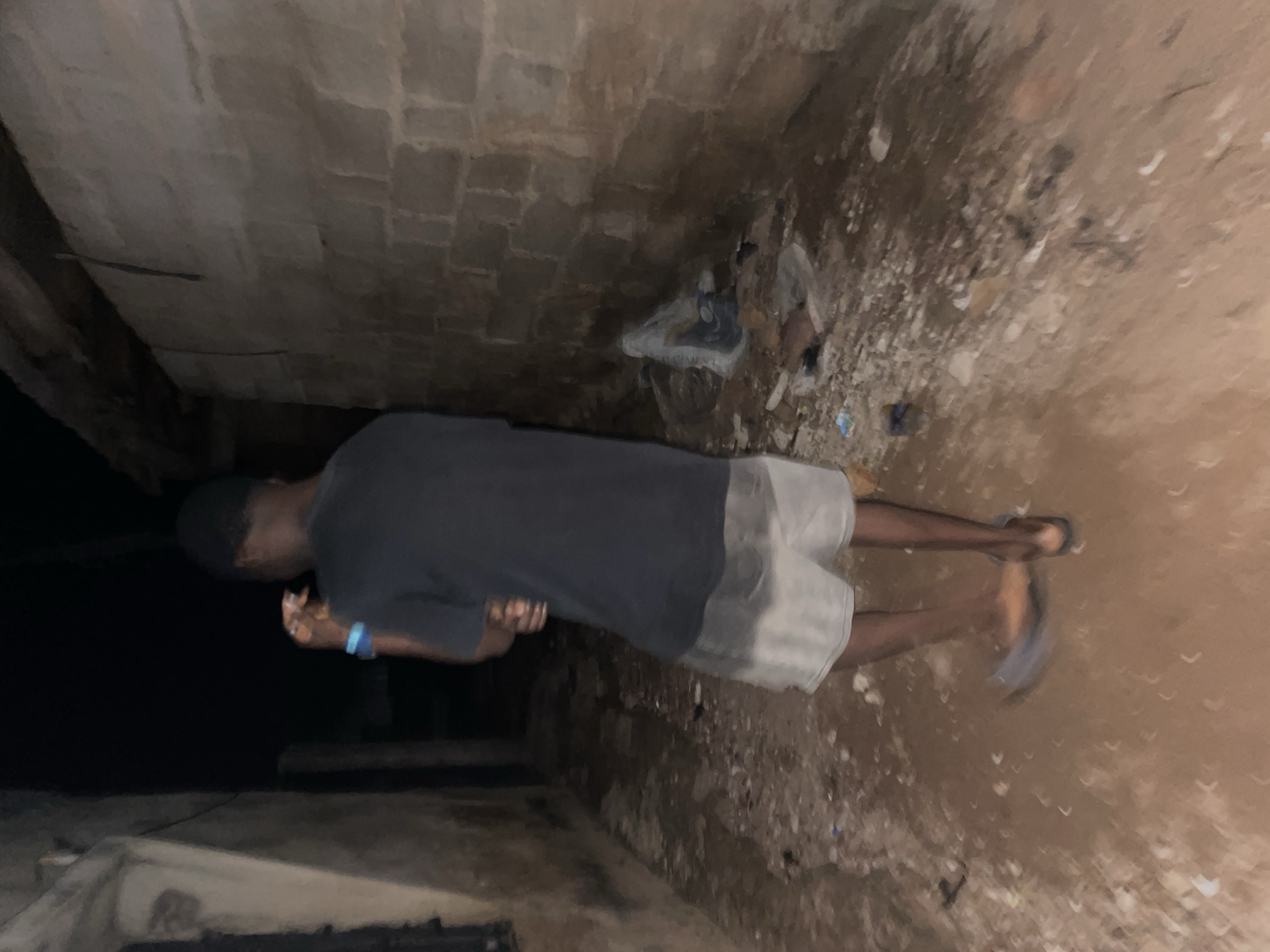 The image depicts a dimly lit alleyway in Lokoja, Nigeria, at night. A person, seen from the back, is standing in the center of the alley. They are wearing a dark t-shirt and light-colored shorts. The ground of the alley is uneven and covered with dirt, pebbles, and scattered debris, including plastic bags. On either side of the alley are rough, textured walls made of concrete blocks. One wall, on the right, appears to be constructed of lighter colored blocks, while the wall on the left is darker and shows signs of wear. The overall impression is one of a neglected urban environment.