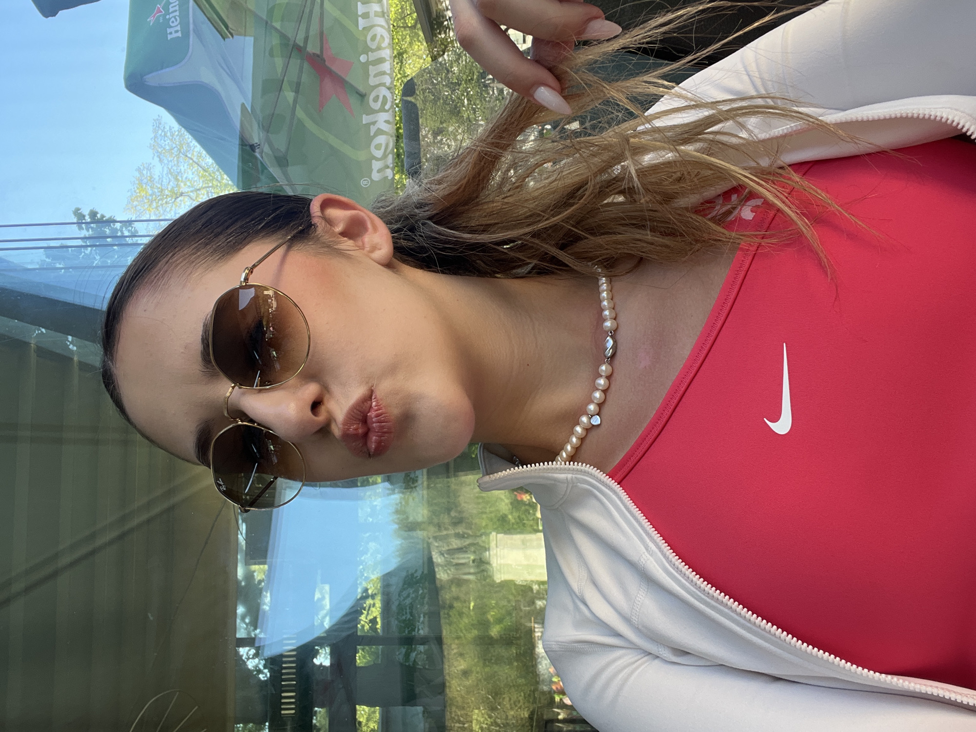 A woman wearing sunglasses, a pearl necklace, a red Nike top, and a white jacket is posing for a photo. She is standing in front of a window, and a Heineken umbrella is visible in the reflection. Without further information, determining the exact location in Blagoevgrad, Bulgaria, is not possible.