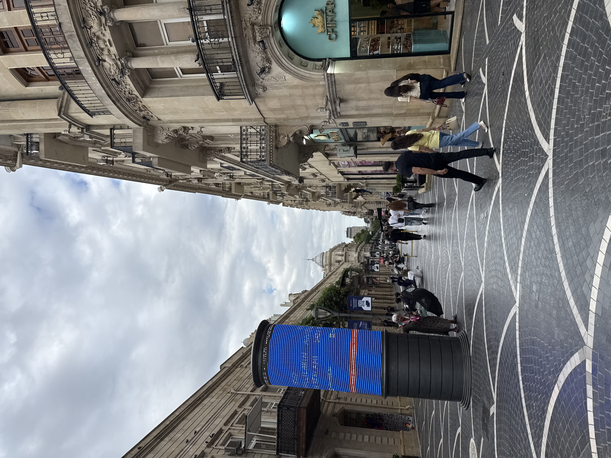 The image captures a street scene in Sabail Raion, Azerbaijan. A cylindrical digital display screen, primarily blue with red and white horizontal lines, stands prominently on the left side of a pedestrianized street. The street is paved with dark gray cobblestones arranged in a pattern of interlocking arcs with white borders. On the right side of the street, a building with ornate stone architecture features multiple balconies with black railings, some of which are adorned with decorative elements and appear to be roosting spots for pigeons. Shops with arched entrances are visible along the street. Several people are walking and sitting on benches, engaged in various activities. A woman in a patterned dress stands near the digital display, holding a black umbrella. Further down the street, people are strolling, some walking in pairs. The sky is overcast with clouds.