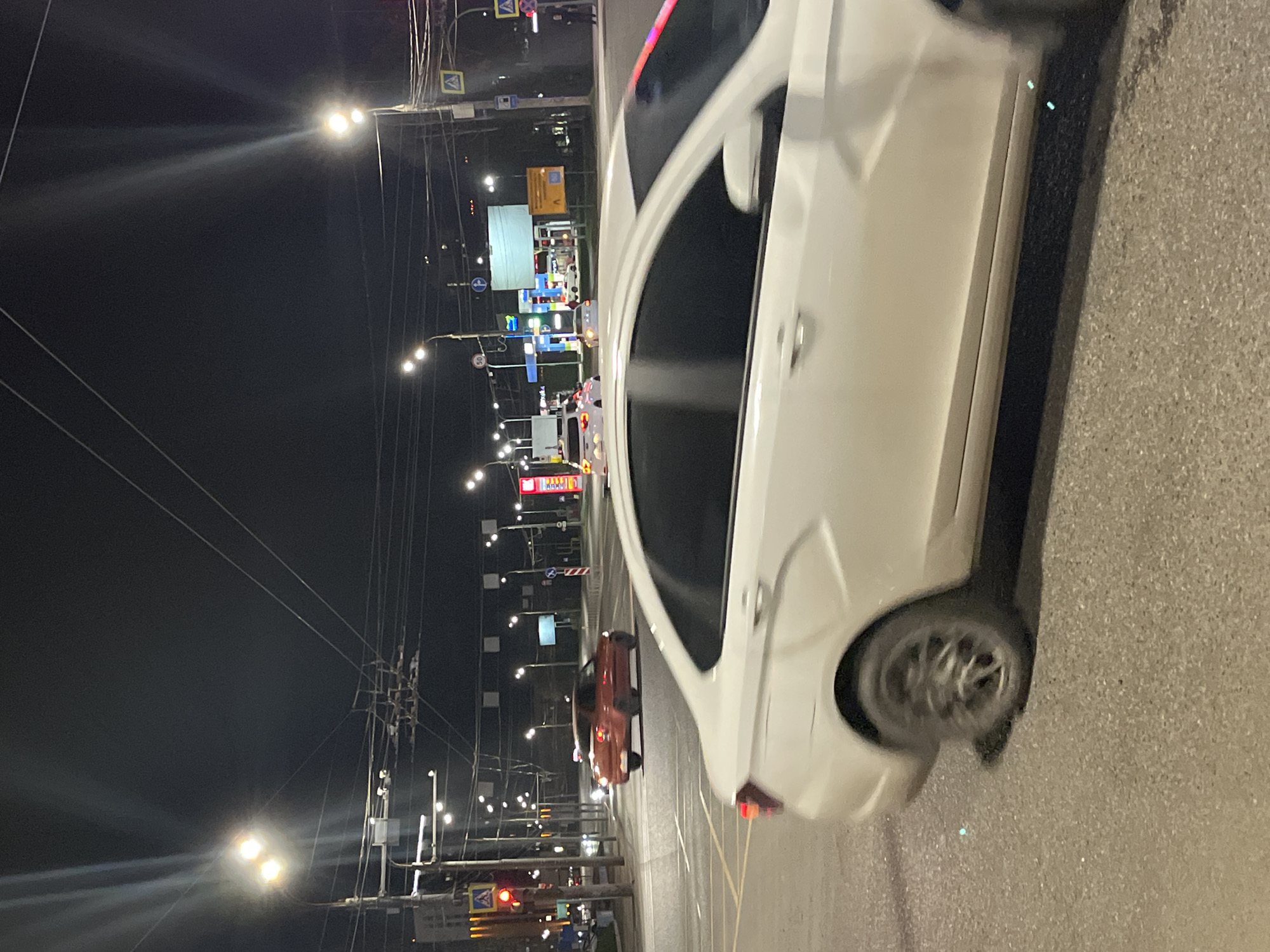 The photograph shows a street scene in Izhevsk, Russia, at night. The scene includes a gas station, street lights, overhead power lines, road signs, and passing cars. A white car is located prominently in the foreground. A red car can be seen in the distance, closer to the gas station. The street appears to be lit by sodium vapor lights, giving the scene a warm, yellow glow.