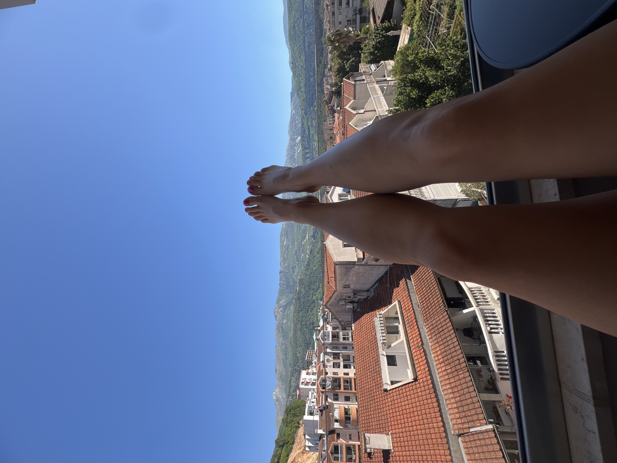 The image shows a first-person perspective from a balcony overlooking the city of Budva, Montenegro. The foreground consists of a person's legs and feet dangling over the balcony edge, with red nail polish on the toes. Below, a cluster of buildings with red-tiled roofs is visible. In the background, mountains stretch across the horizon under a clear blue sky. The scene suggests a sunny day and a relaxed atmosphere.