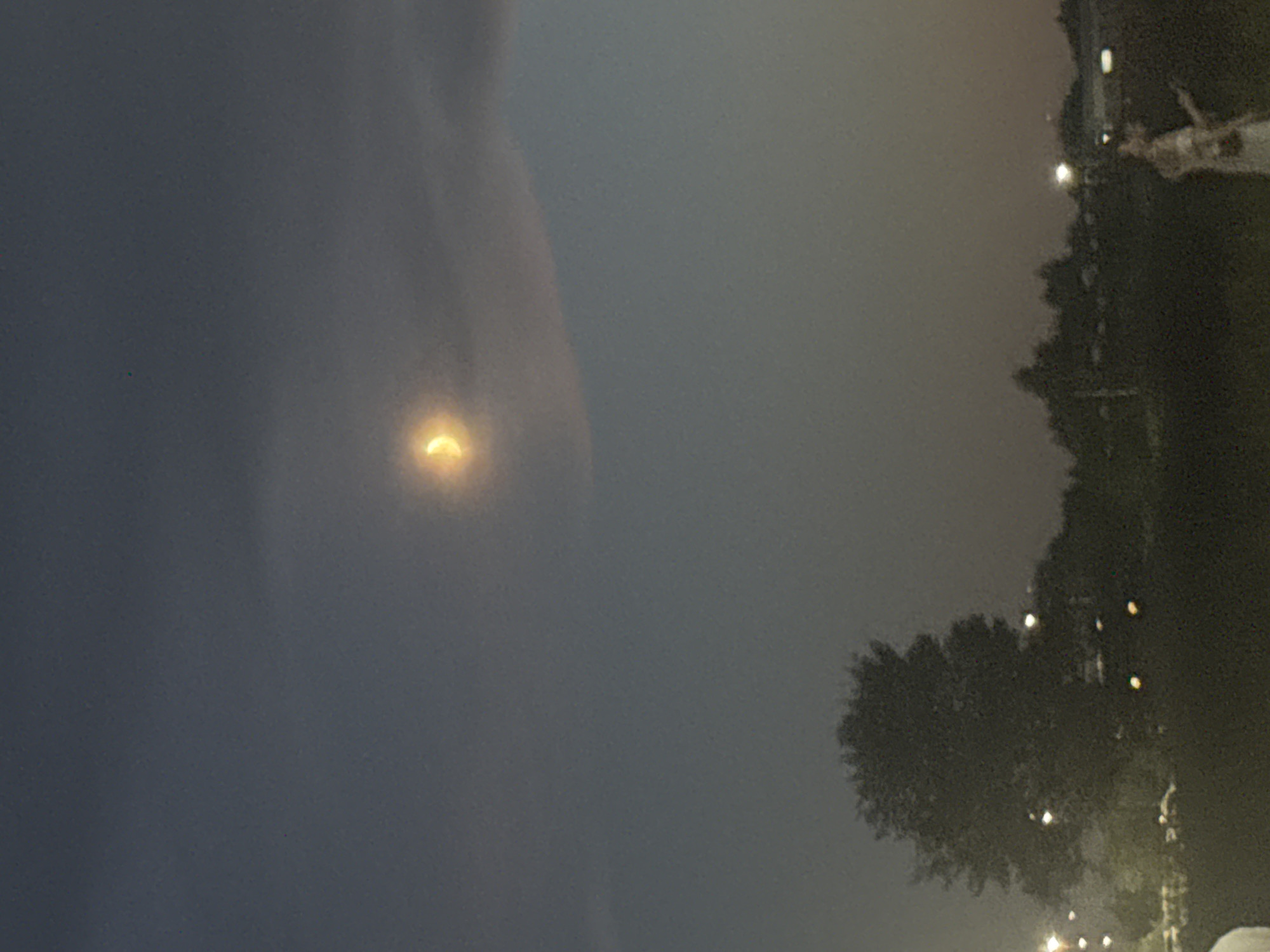 The image depicts a dimly lit outdoor scene at dusk or night, with a crescent moon visible in the upper center of the frame. The sky is a gradient of dark blue to a lighter, hazy gray. In the foreground, silhouetted trees and a person in a light-colored dress are visible. Distant lights suggest an urban or suburban environment. The overall impression is one of a serene, twilight landscape. The location is Komsomolsk-on-Amur, Russia.