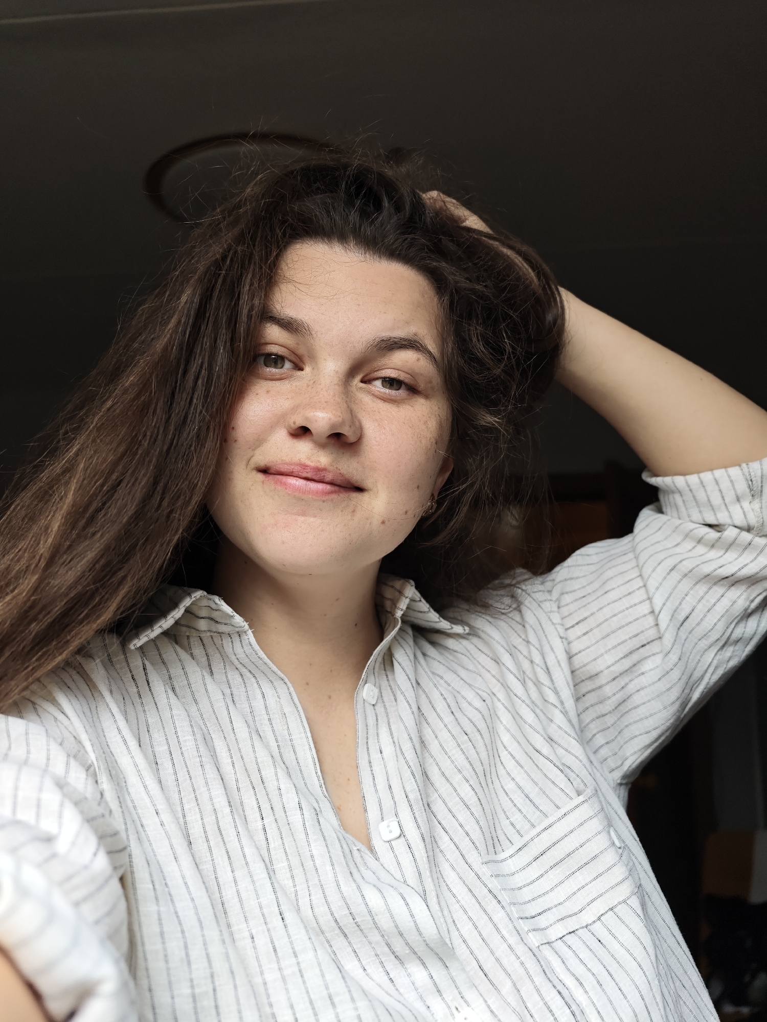 A young woman with light skin, brown hair, and freckles is posing for a selfie indoors. She is wearing a white and gray striped button-down shirt. Her right arm is raised, with her hand partially in her hair. The background is dark, with a hint of a light fixture above her head. The location cannot be determined as Chaikovsky, Russia, from the image.