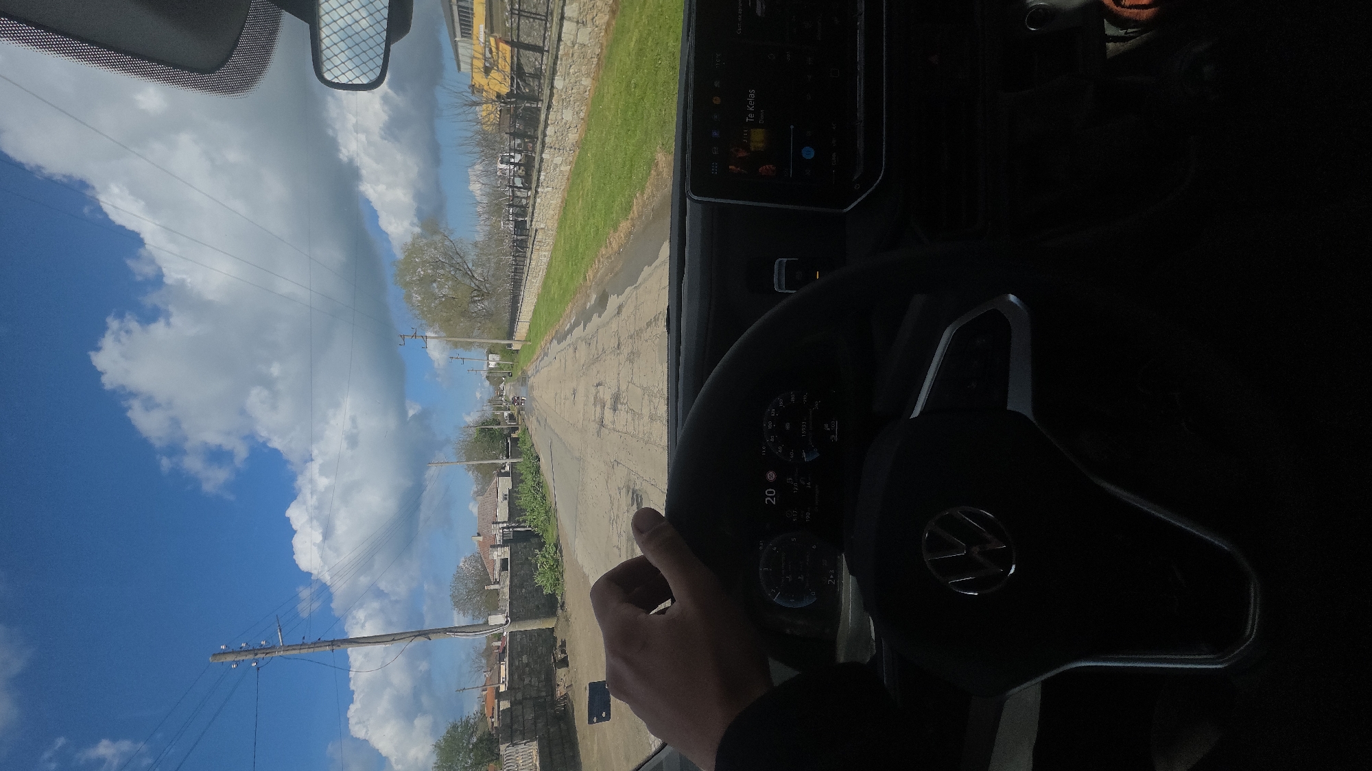 A person is driving a Volkswagen vehicle on a cracked asphalt road in Balgarevo, Bulgaria. The view through the windshield shows a street lined with houses and telephone poles under a blue, cloudy sky. The car's dashboard and steering wheel are visible, and a hand is seen holding the steering wheel. The navigation screen displays "Te Kelas" by Coke Studio. The environment appears rural or suburban.
