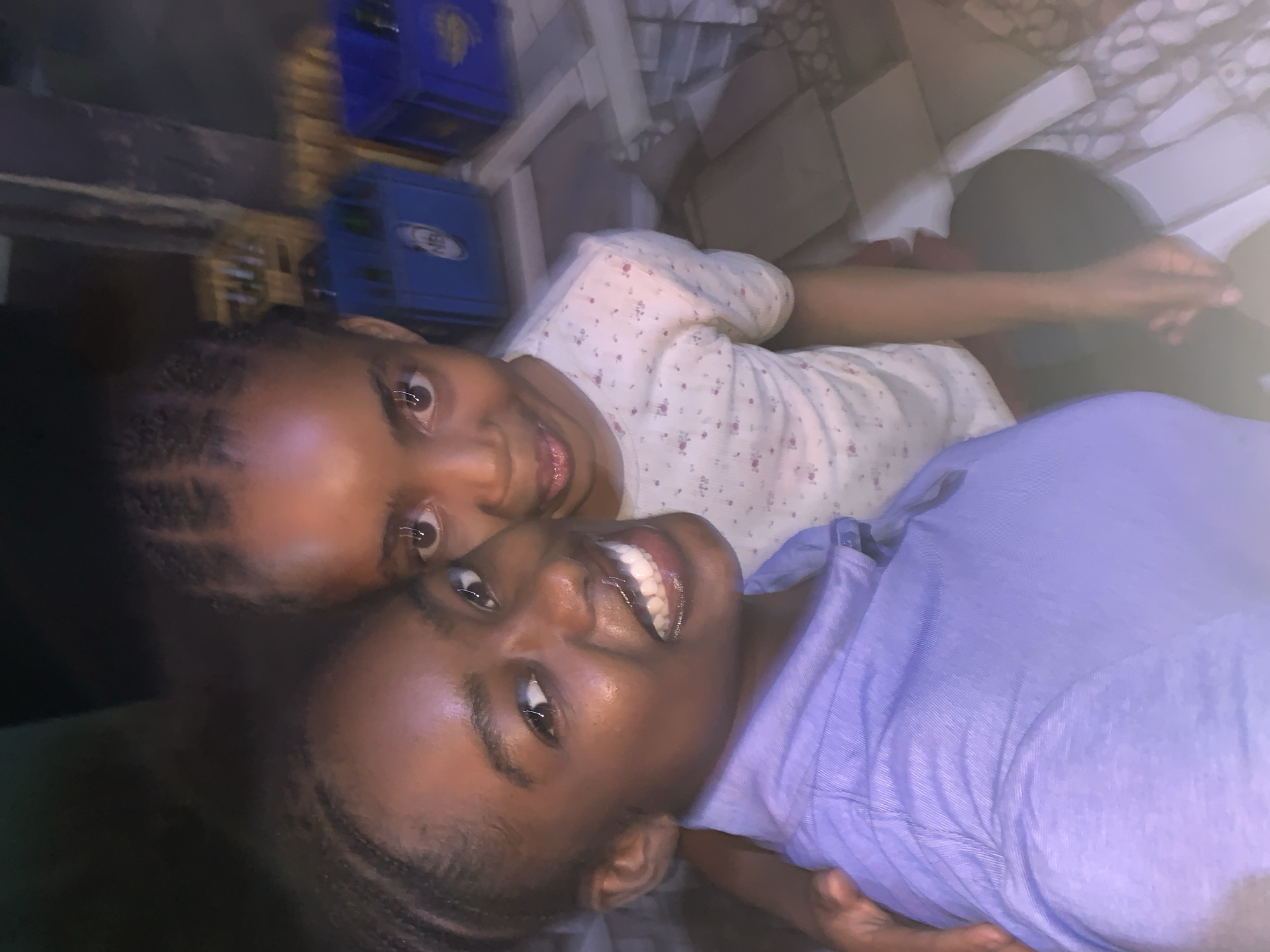 Two young girls are lying down together indoors. One is wearing a light purple t-shirt, and the other is wearing a white shirt with a floral pattern. They are both smiling at the camera. The location is New Stadium, Nigeria. There are blue crates in the background, along with flooring that appears to be tiled or otherwise textured. They appear to be posing for a photograph.