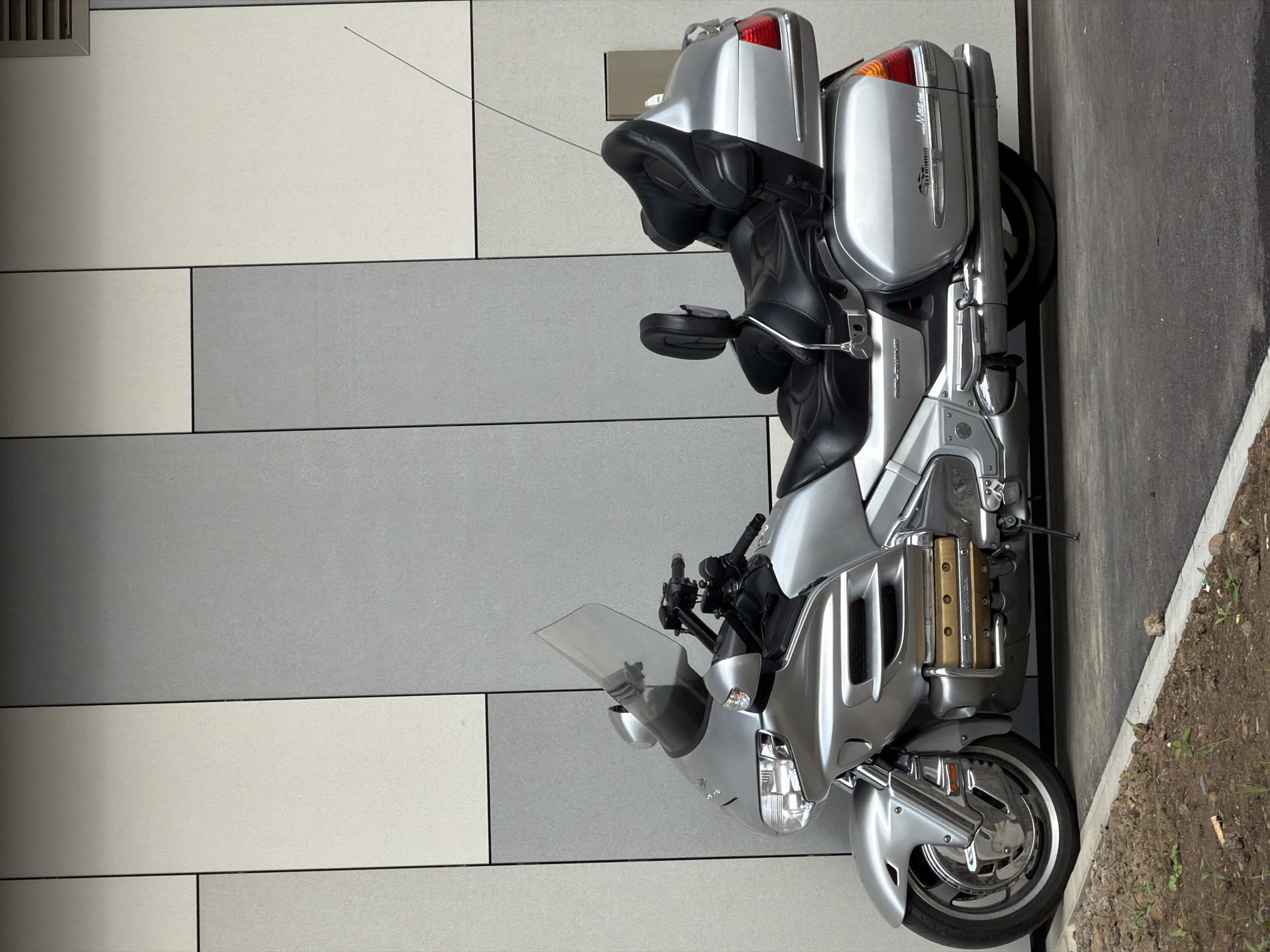 A silver Honda Gold Wing touring motorcycle is parked beside a building in Esch-sur-Alzette, Luxembourg. The building's facade features alternating horizontal rows of light gray, gray, and off-white rectangular panels. The motorcycle is parked on asphalt pavement next to a narrow strip of soil with sparse green vegetation. The motorcycle is oriented with its front towards the bottom of the frame, showcasing its windscreen, handlebars, and storage compartments. The rear of the motorcycle includes tail lights and passenger seating. The scene appears to be a static shot of the parked vehicle.