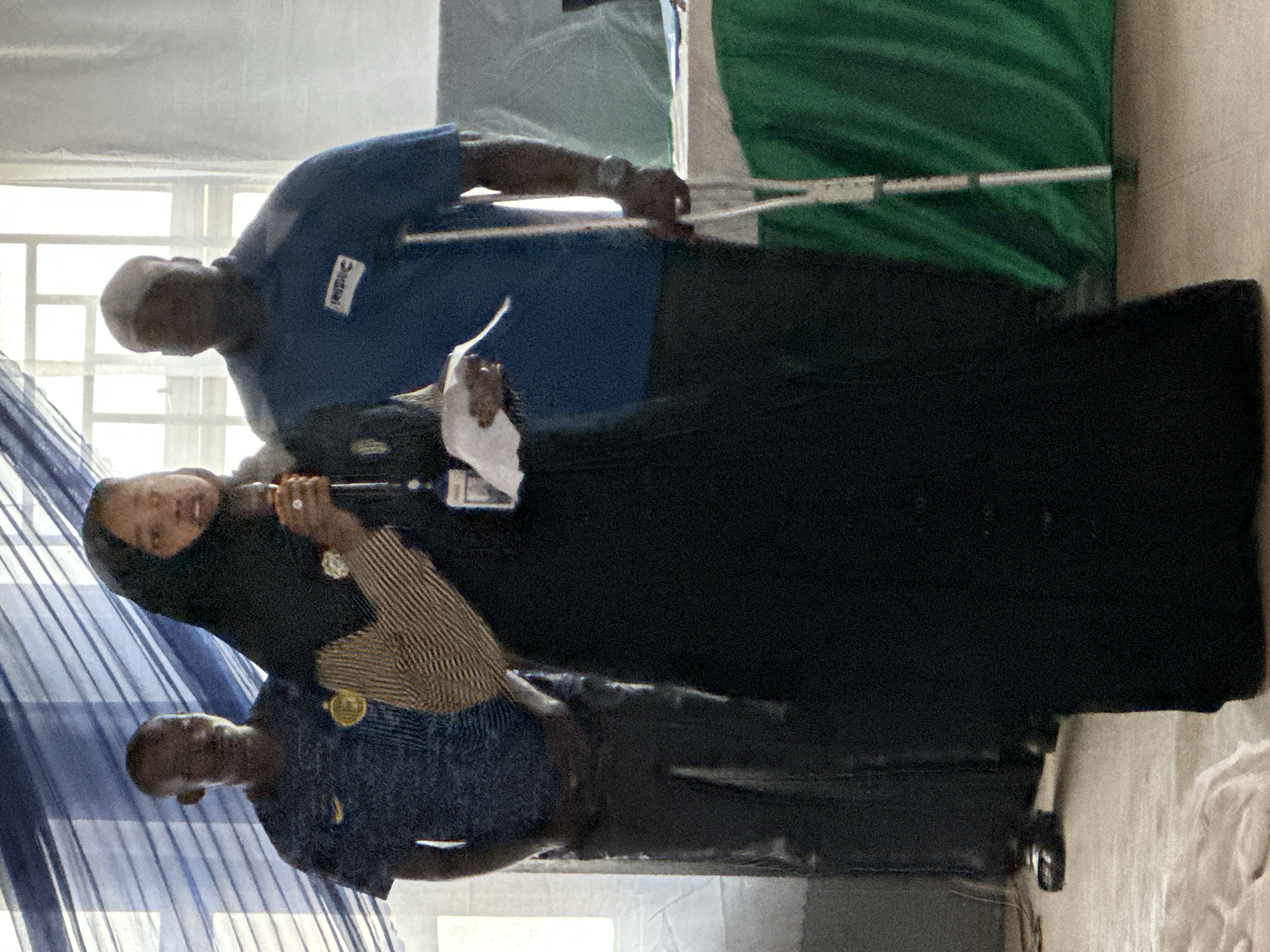 The image shows three individuals standing indoors, likely at an event or gathering in Unguwan Dosa, Nigeria. The setting appears to be a room with natural light coming from windows partially covered by blue striped curtains or fabric on the left side.
In the foreground, a woman wearing a black hijab and a striped top is centrally positioned. She holds what appears to be a microphone or a similar handheld device in her right hand, suggesting she might be speaking or presenting. A ring is visible on her finger. To her right stands a man with a bald head, dressed in a blue polo shirt that features a white rectangular logo on the chest, possibly reading "Victor." He is holding a long, thin white pole or stick in his right hand and appears to be holding a small white document or paper in his left hand, near the woman. To the woman's left, another man with a bald head is visible, wearing a dark blue or black shirt with a round gold badge or logo on the chest.
The background includes a large, dark-colored structure, possibly a display stand or backdrop. Further to the right, a significant portion of green fabric is visible, draped and folded, strongly suggesting it is part of a national flag, such as the Nigerian flag, given the context. The overall atmosphere appears semi-formal, indicative of a public event or presentation. The image has a slightly blurry quality with some visual noise.