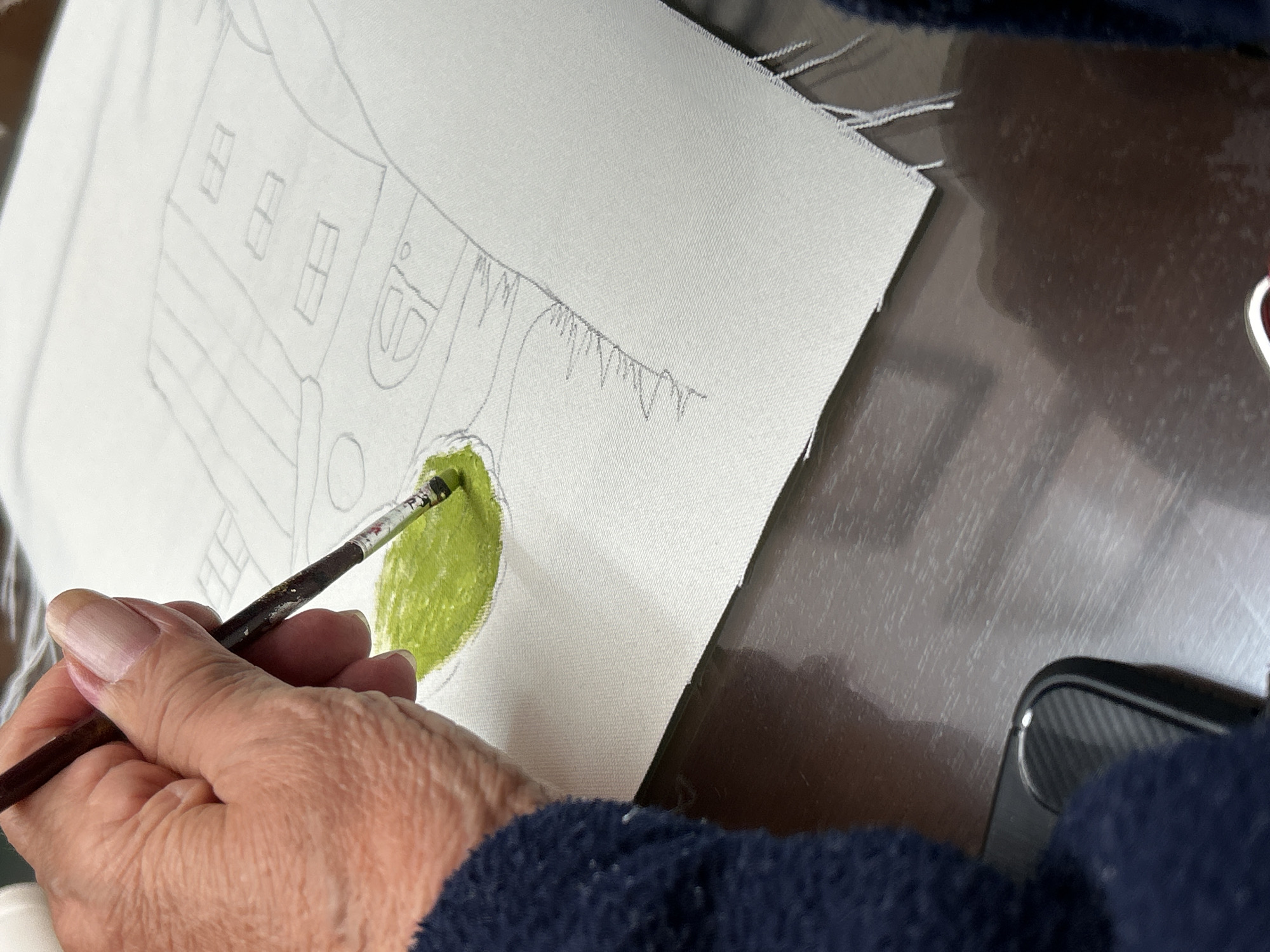 A close-up shot shows a person's hand holding a paintbrush, applying green paint to a canvas. The canvas is stretched on a frame and has a pencil sketch of a building and foliage. The person is wearing a dark blue sweater. The background includes a glossy surface and an object (possibly a phone) with a textured back. The location is undetermined.