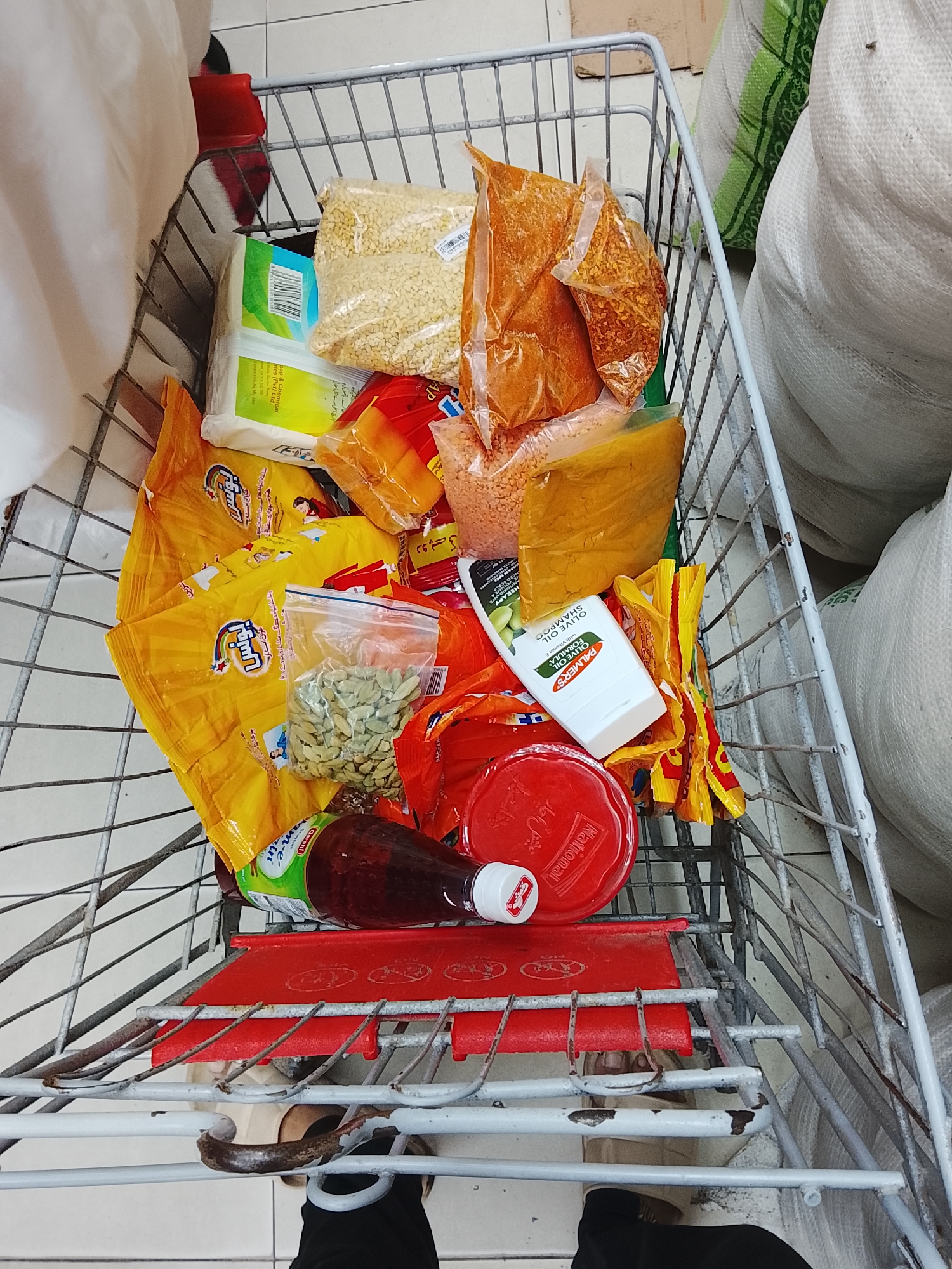 The image shows a shopping cart filled with grocery items. These include bags of lentils, spices (such as chili flakes and cardamom), turmeric, a bottle of olive oil shampoo (Palmer's), tomato ketchup, and yellow-packaged goods. The cart is made of metal and has a red plastic seat. The shopper's legs and feet are visible at the bottom of the frame. Bags of unidentifiable goods are on either side of the cart. The image was taken in the city of Gulgasht Colony, Pakistan.