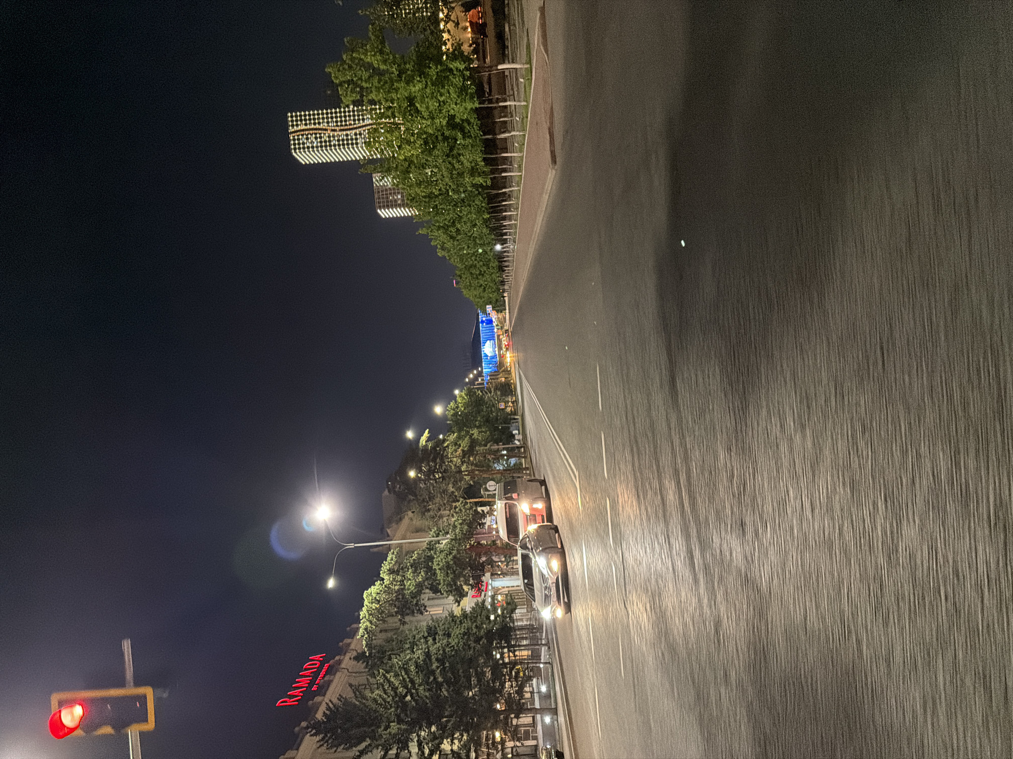 The image captures a night scene on a city street in Astana, Kazakhstan. In the foreground, the road surface is visible with motion blur, indicating the photo was taken from a moving vehicle. Two cars are driving on the road; a silver sedan is in the left lane, and a white minivan is in the right lane. Streetlights illuminate the scene, and trees line the right side of the road, with modern buildings in the background. A red traffic light is visible on the left side of the frame, and a sign with the word "RAMADA" is displayed on a building. The overall impression is of a well-lit urban environment at night.
