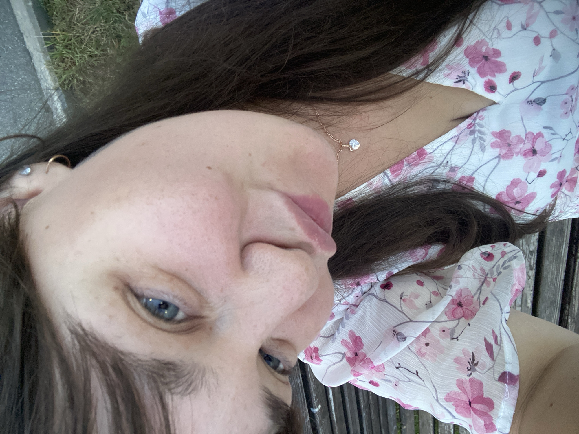 The image is a close-up selfie of a woman with long, dark brown hair and blue eyes. She is wearing a white dress with a pink floral pattern and a delicate necklace with a heart-shaped pendant. The woman is leaning forward, with her face partially obscured by her hair. The background shows a hint of greenery and a wooden bench, suggesting an outdoor setting, possibly a park or garden. The image is taken outdoors during the daytime. No specific location in Votkinsk, Russia is identifiable from the image alone.