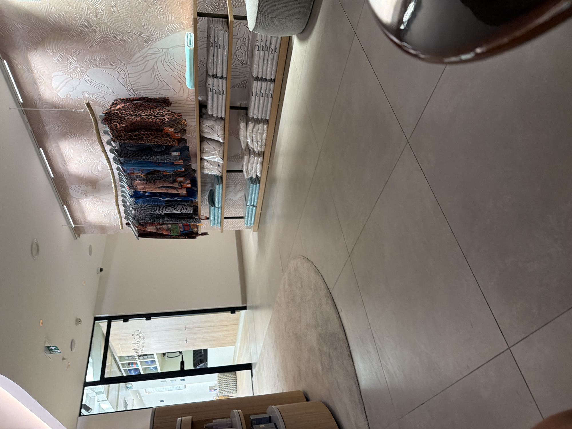 The image depicts the interior of a retail store. On the left, a rounded wooden display unit with shelves holds various products. Adjacent to it is a glass-paned door with a sign that reads "Absolute Studio." Inside the doorway, more shelves are visible, some containing blue spherical objects. The floor is tiled in a neutral tone.
To the right, a clothing rack is suspended from a branch-like fixture, displaying an assortment of colorful garments, including a leopard-print shirt. Behind the clothing rack is a wall with a patterned wallpaper featuring white line drawings of figures and abstract shapes. Below the clothing rack are wooden shelves displaying stacks of neatly folded items, many of which appear to be textiles, possibly towels or linens, wrapped in plastic. A light blue folded item is placed on the top shelf. A round, ribbed, light grey ottoman is positioned in the far right of the frame. The lighting is soft and ambient. The location is not identifiable as Boreti, Montenegro based on the visual information present in the image.