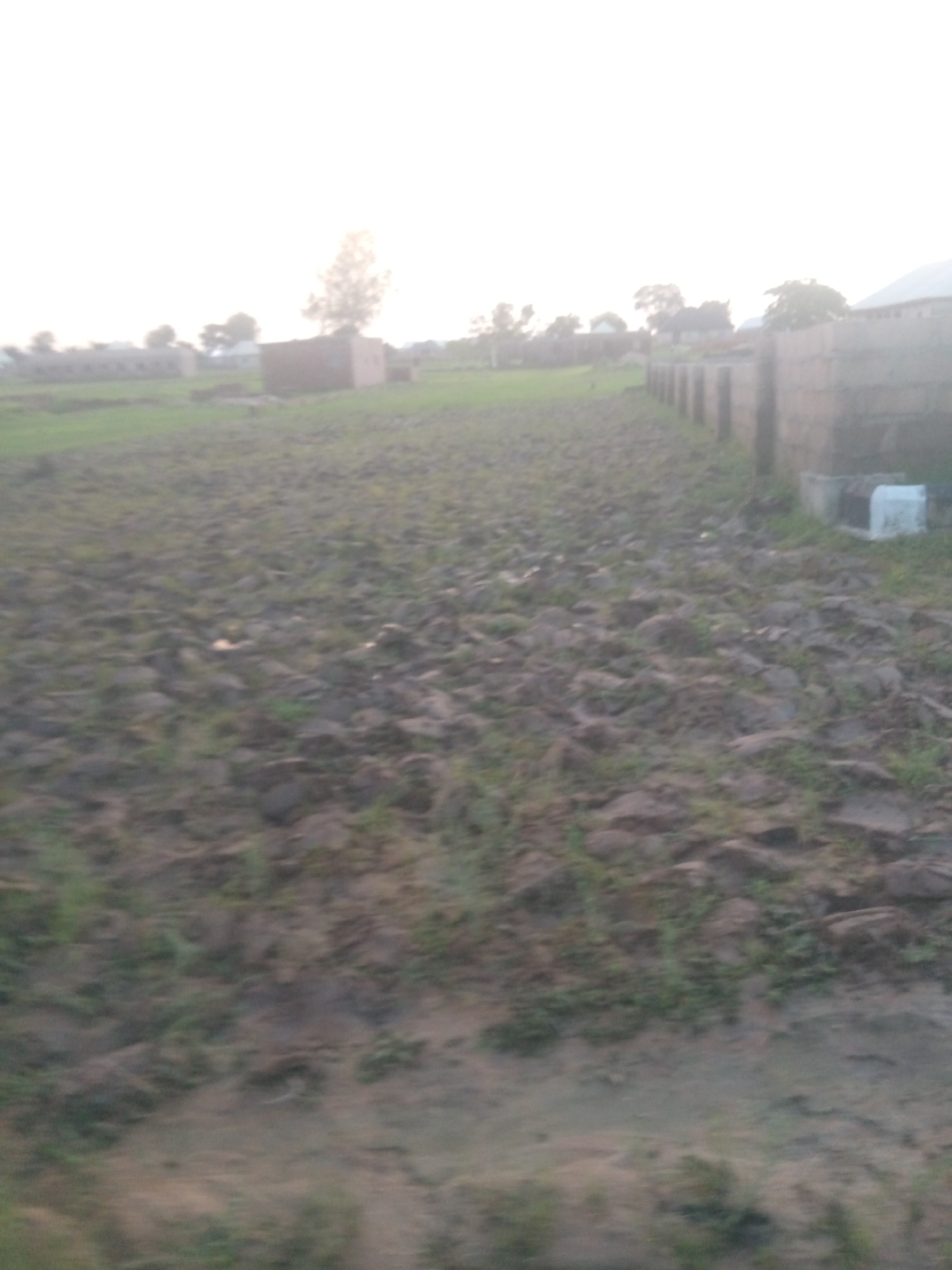 The image depicts an outdoor scene in Jalingo, Nigeria. The foreground is a field of tilled earth with scattered rocks and sparse vegetation. A dirt path runs alongside a brick wall on the right side of the frame. In the background, there are several buildings, including what appears to be a school or administrative building with a long, rectangular structure. A smaller, solitary brick building is also visible. Trees and other vegetation dot the landscape, and the sky is overcast with a bright, hazy sun. The overall impression is one of a rural or semi-urban environment.