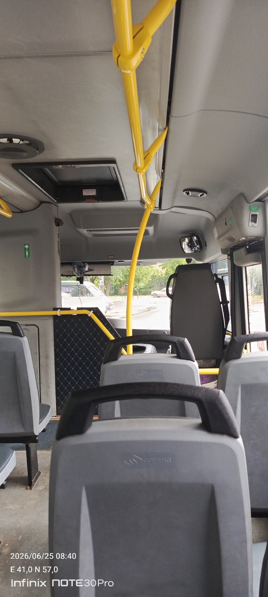 This is a photograph taken inside a public bus, looking towards the front. The interior is characterized by gray plastic seats with black handles and yellow grab poles mounted on the ceiling and along the sides. A patterned black panel is visible on the left side wall. Through the windows, a street scene with trees and a light-colored van is discernible. The timestamp in the bottom left corner indicates the photo was taken on June 25, 2026, at 08:40, with GPS coordinates E 41.0 N 57.0, suggesting the location is within the city of Ivanovo, Russia. The phone model "Infinix NOTE 30 Pro" is also imprinted on the image. There are no people visible in the frame.