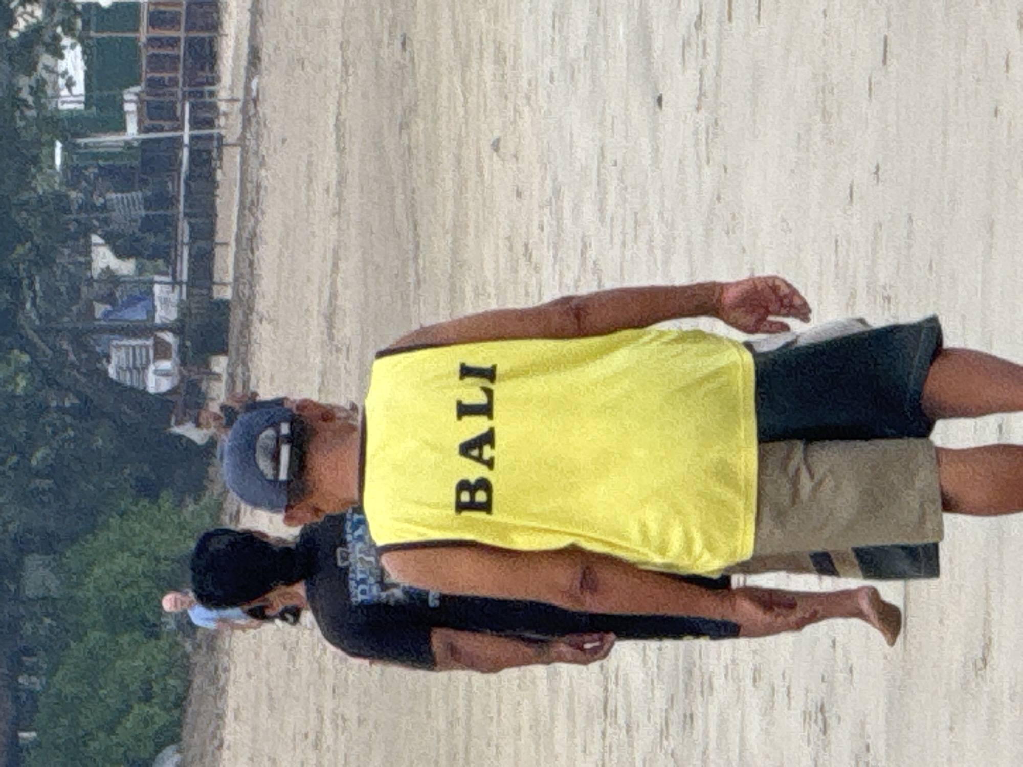 On a sandy beach in Jimbaran, Indonesia, a man in a yellow "BALI" tank top and dark shorts stands barefoot. Another person in a dark shirt stands nearby, also barefoot. In the background, trees and buildings are visible.